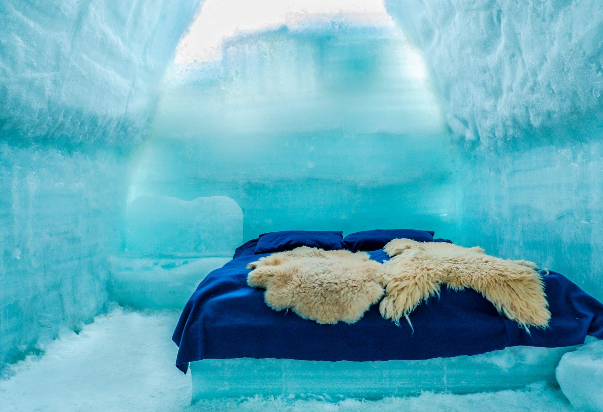 A bed inside an igloo with two beige furs, blue flat sheets and pillows.