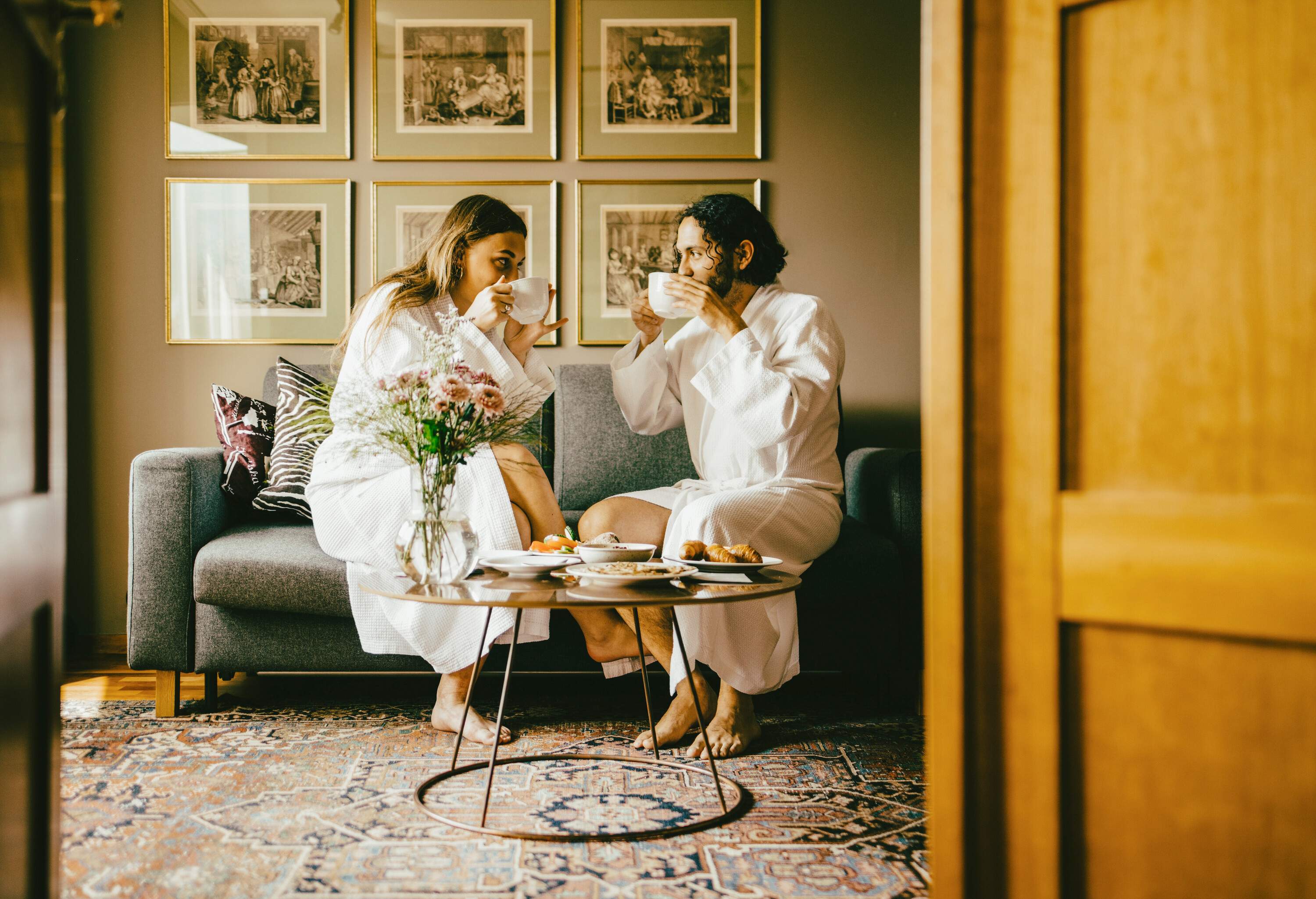 A couple in their white bathrobes enjoying their drinks in large white mugs as they sit on a sofa facing each other.