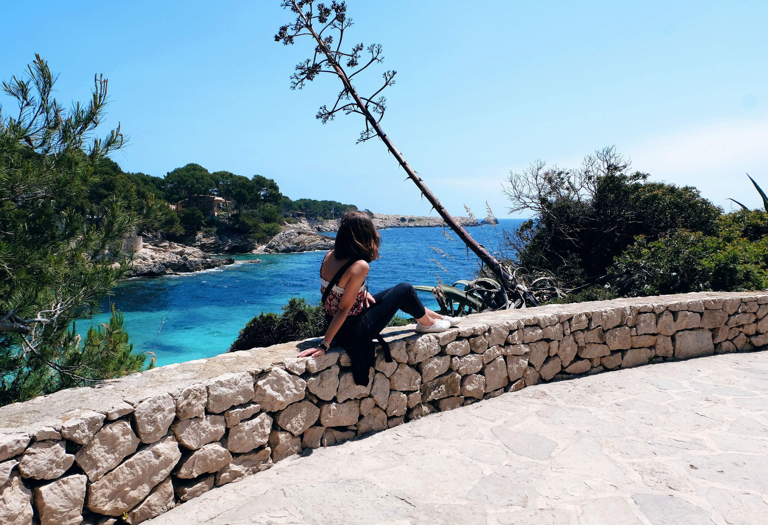 A woman sitting on a low wall along a road, staring out at the gorgeous blue sea.