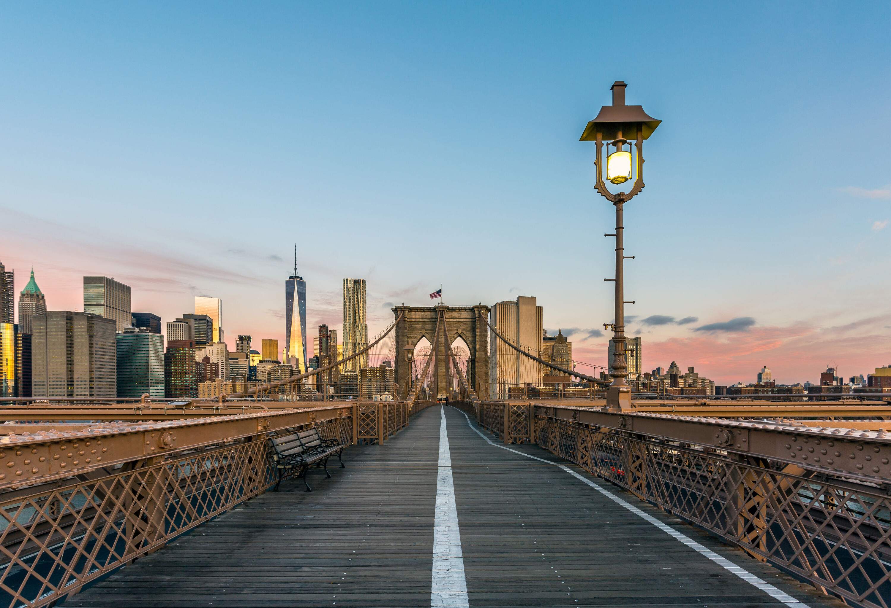 A relatively empty Brooklyn Bridge with a bench on the side and lofty skyscrapers in the surrounding area.