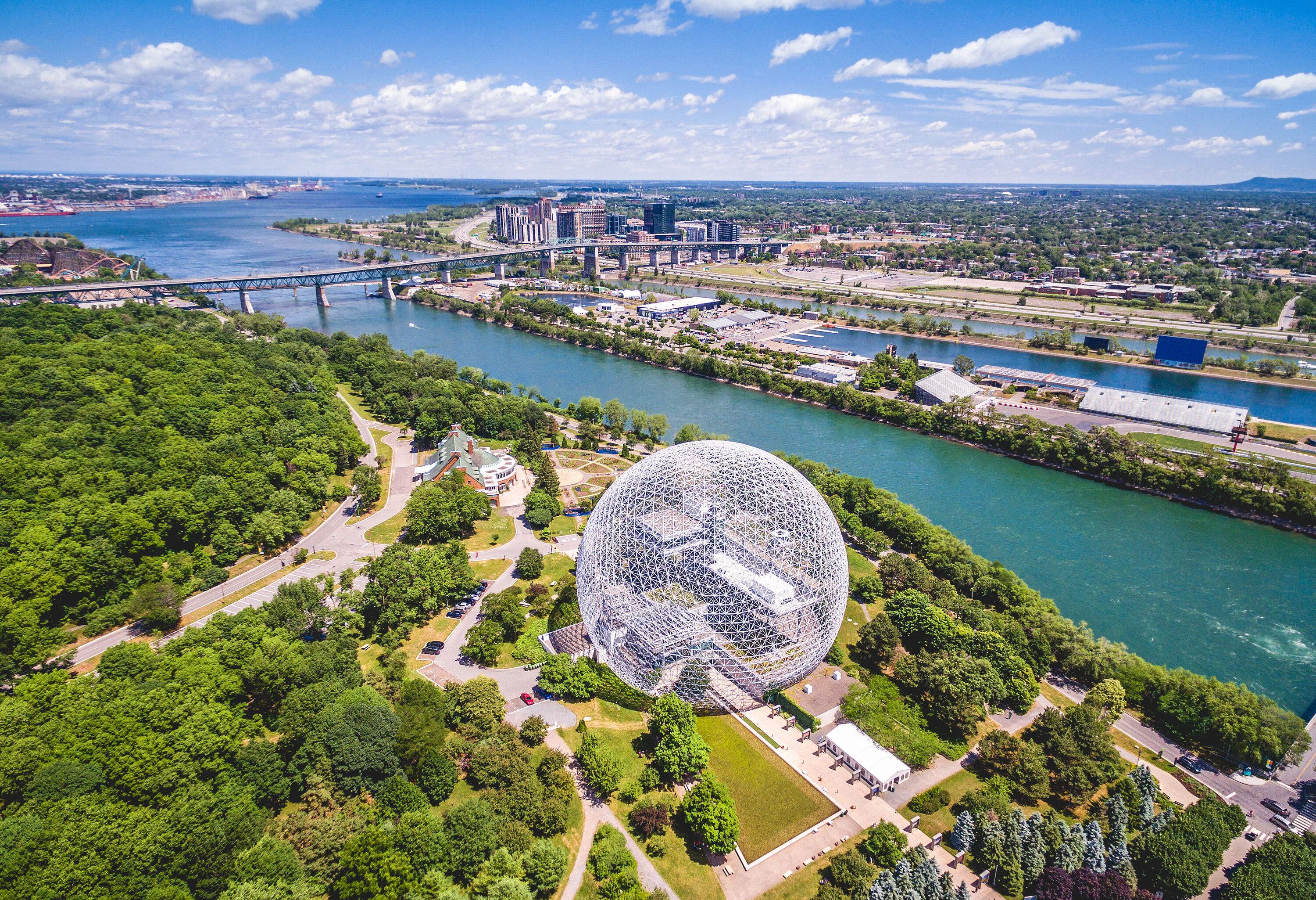 A spectacular geodesic dome museum nestled in a lush green park alongside the river spanned by a long bridge.