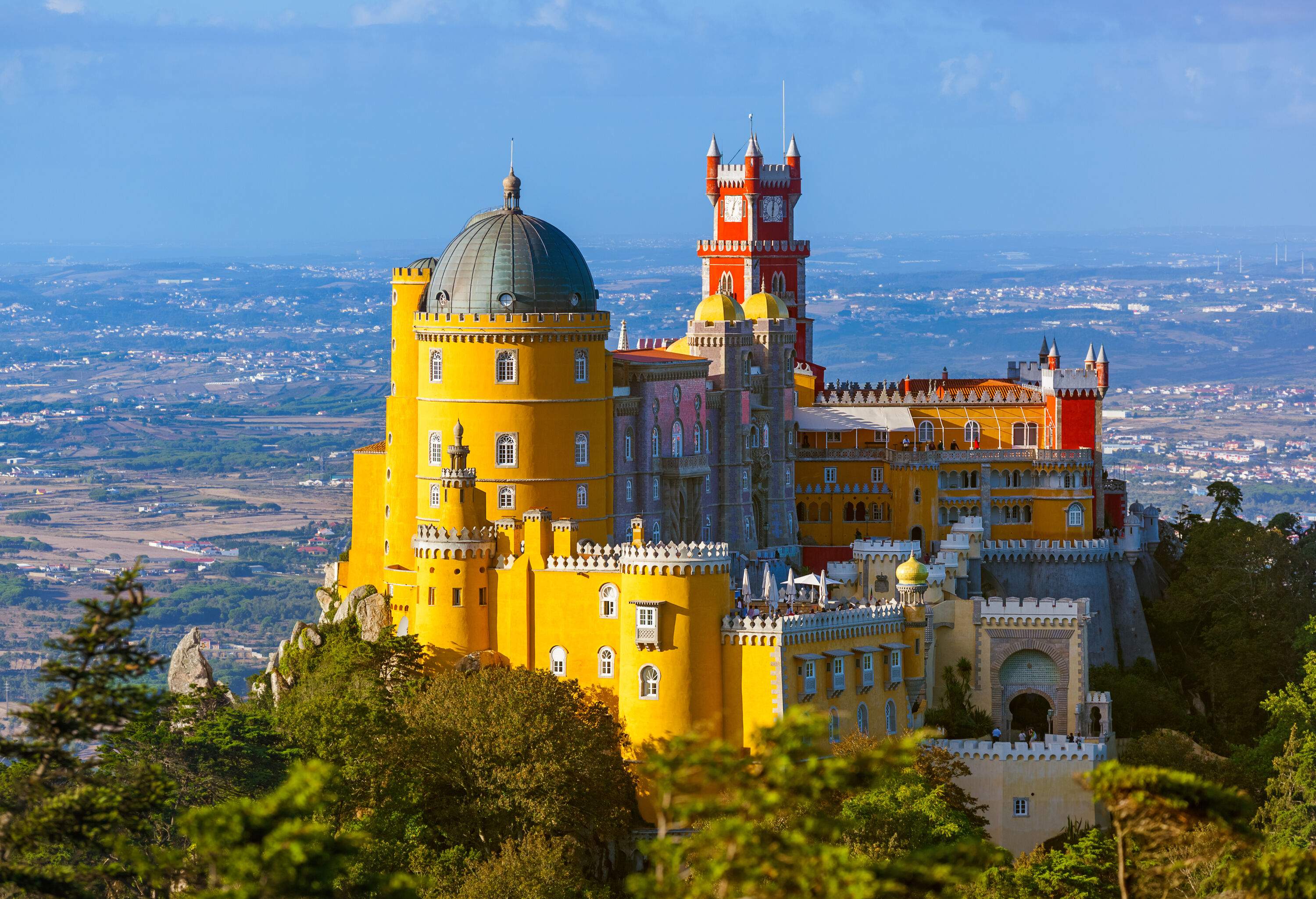 A vibrant, colourful Romanticist castle perched on a hilltop overlooking a vast land sprinkled with houses.