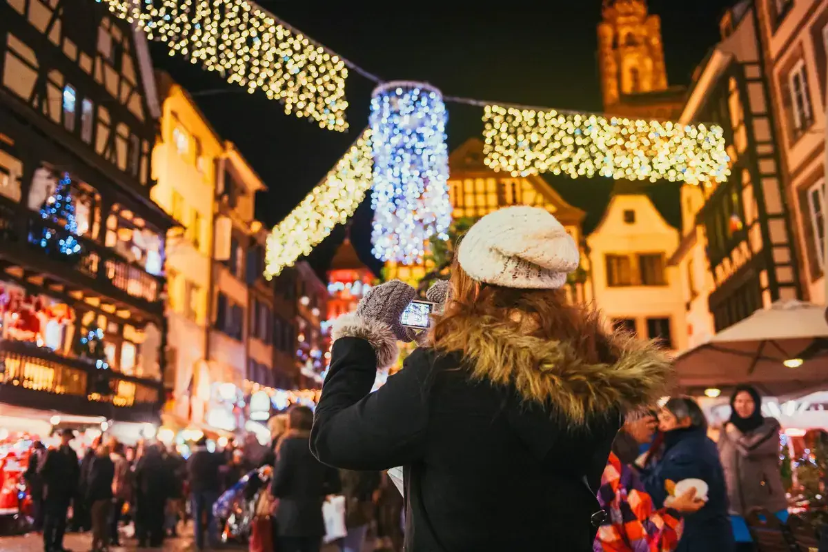 A woman wrapped up in winter clothes taking a photo of Christmas lights at a Christmas market in France