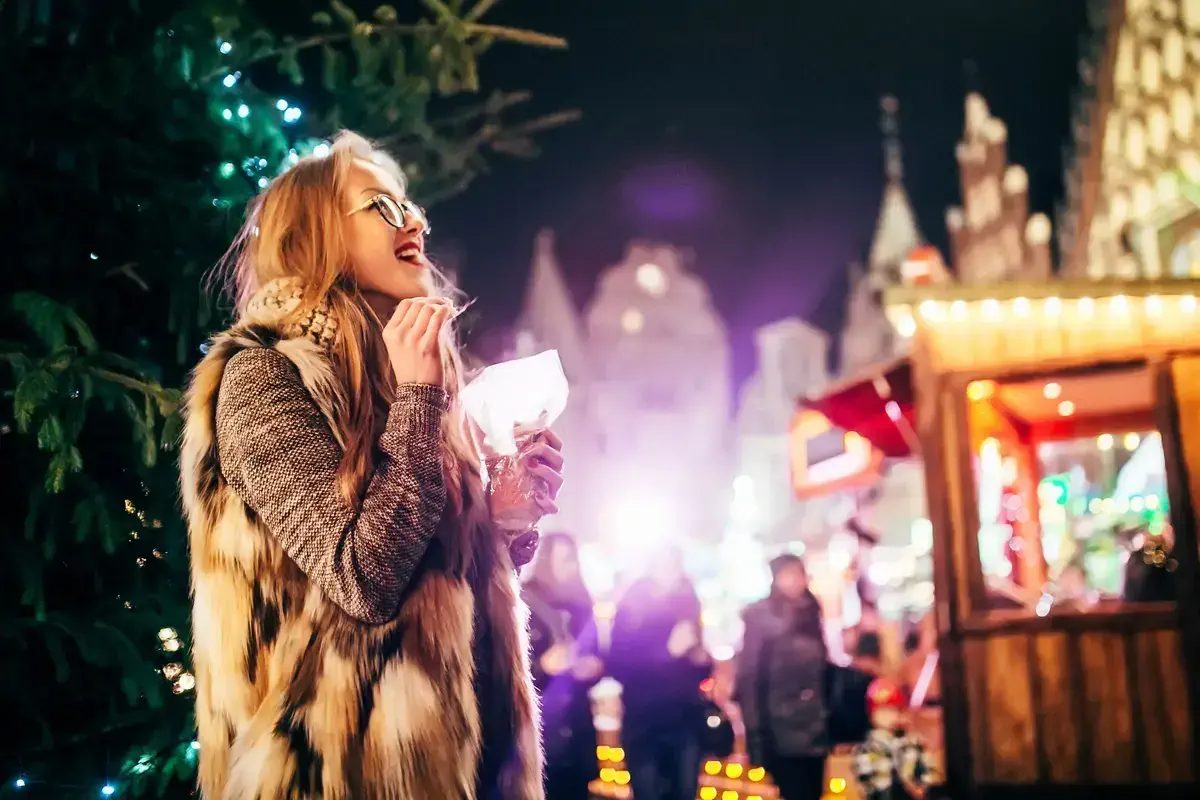 A happy woman with glasses and blonde hair eating a festive snack at a Christmas Market in France
