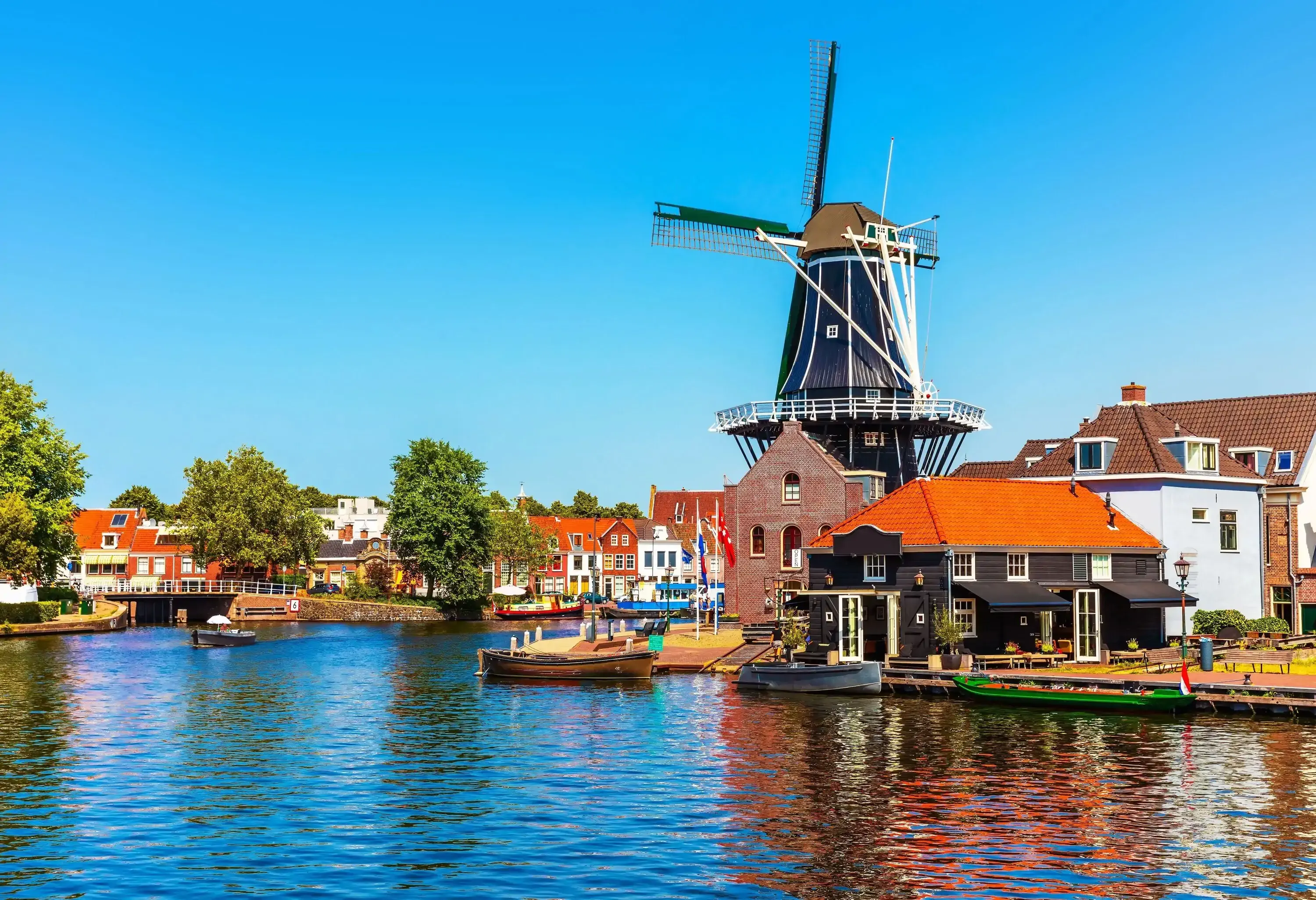 A navy blue windmill rises above a cluster of waterfront homes along a water canal with anchored boats.