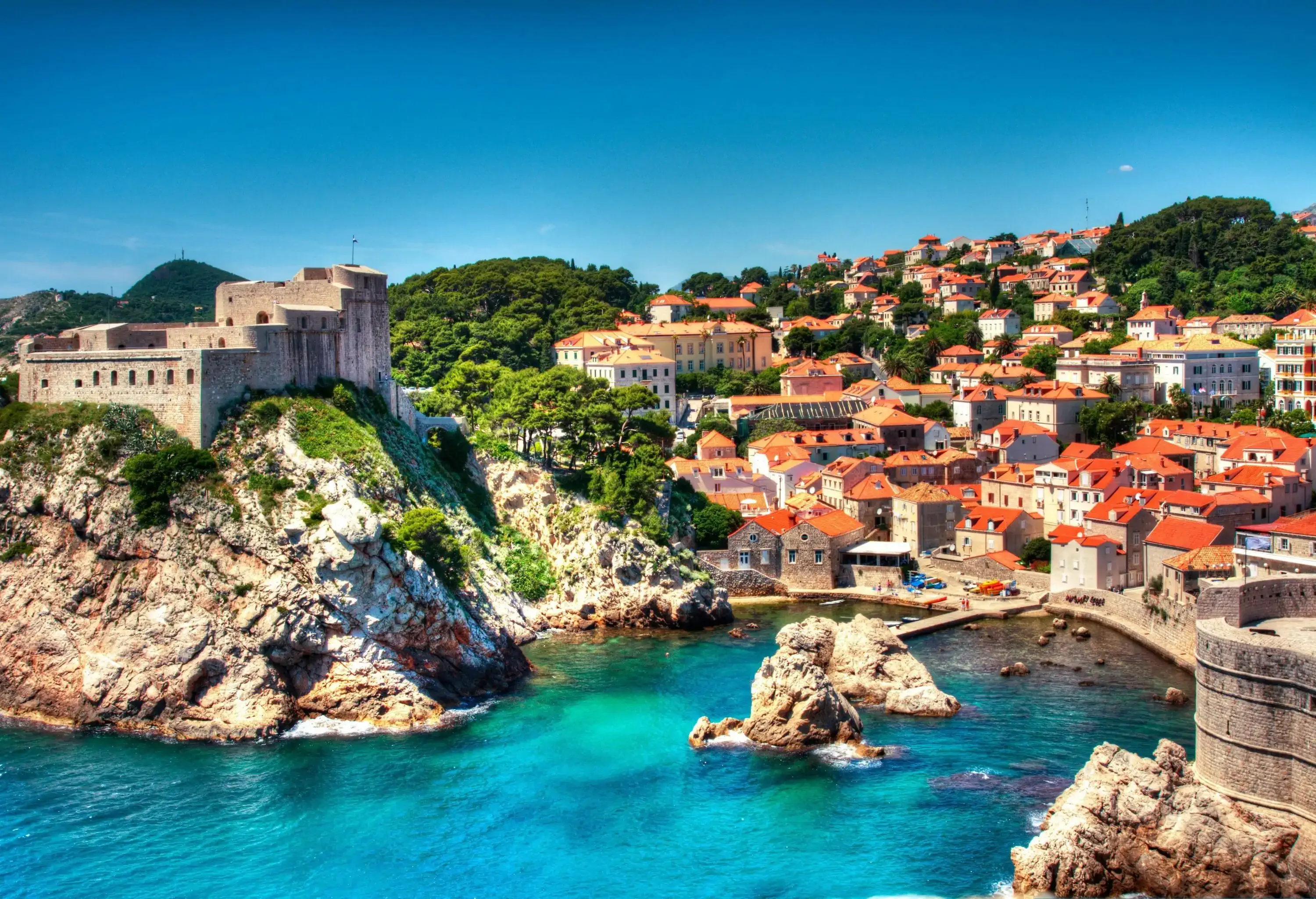 A triangular fortress crowned on a rock cliff behind houses layered on the hill surrounded by the blue-green sea.