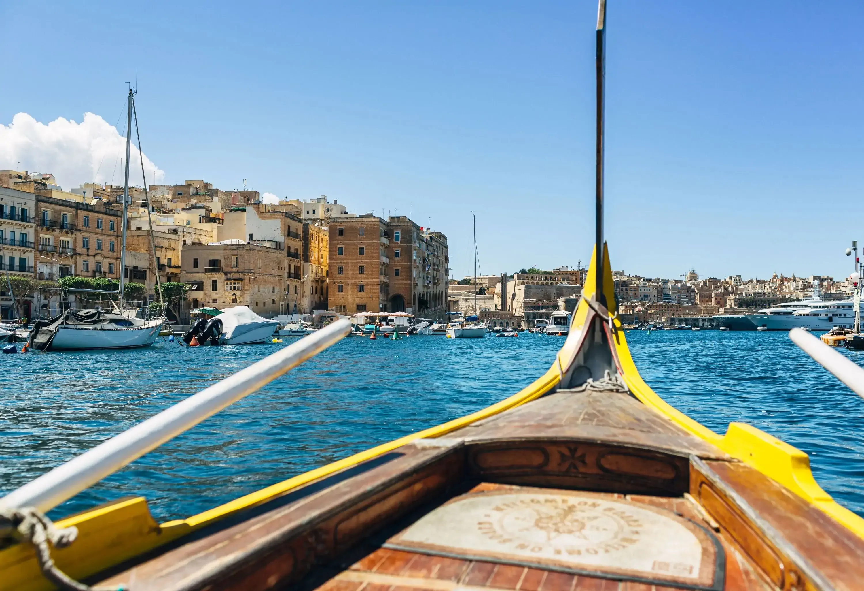 A wooden boat floating in the sea with views of boats in the harbour and hillside buildings on the coast.