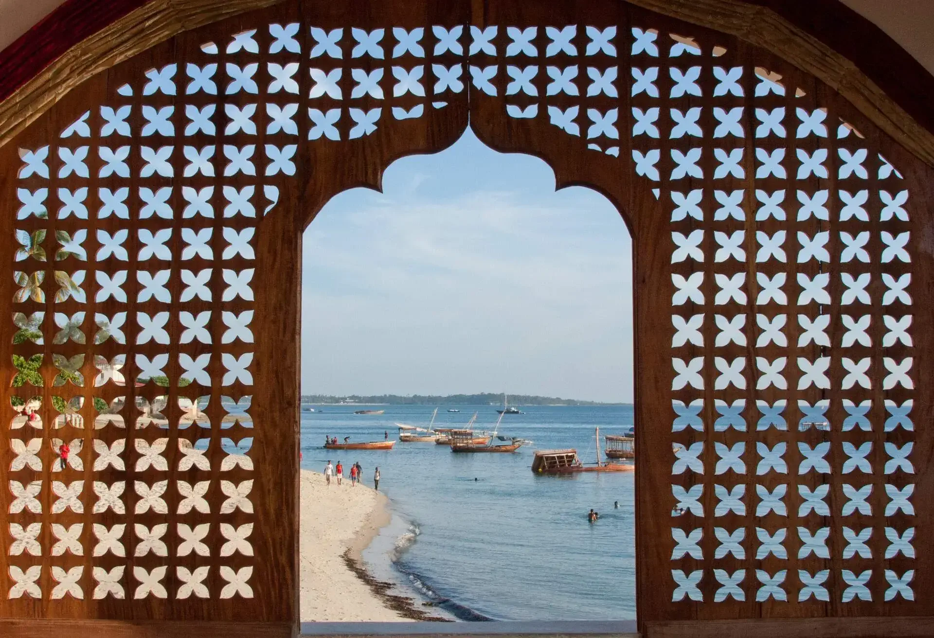 A view of Stonetown beach and bay through a lattice work window of a hotel.. There are shipwrecks in the background and people walking along the beach. Stonetown, Zanzibar, Tanzania, East Africa.