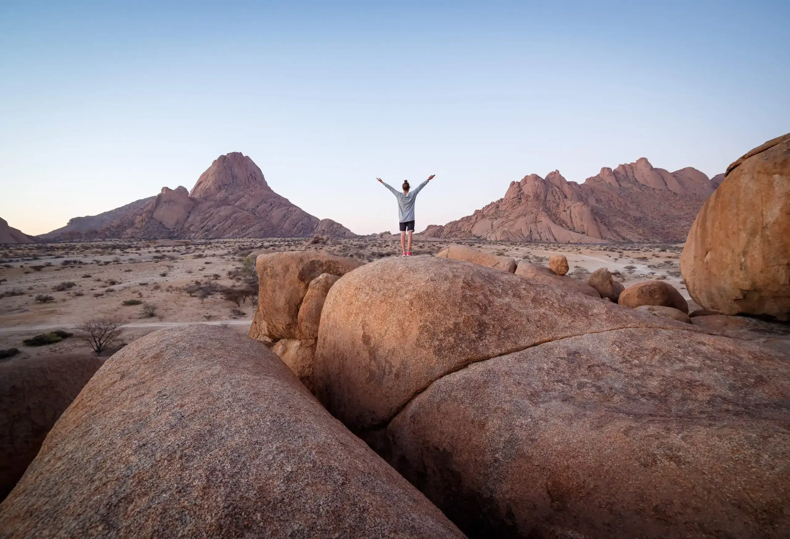 A woman standing on a big rock in the mountains with both arms raised up high.