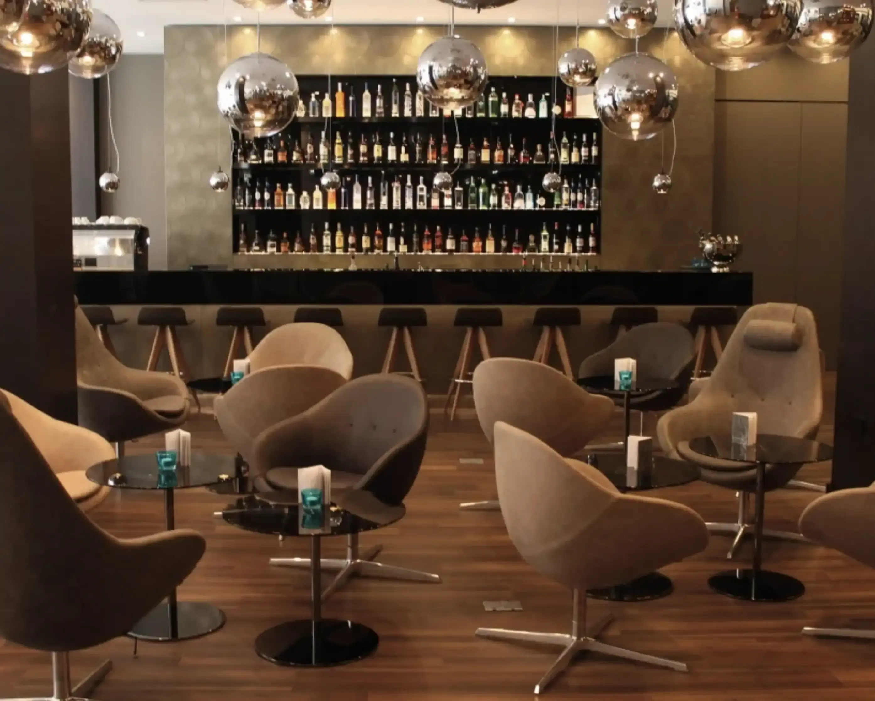 Modern lounge bar with brown armchairs, round tables, and a sleek bar stocked with bottles under soft lighting. Chrome pendant lights hang above.