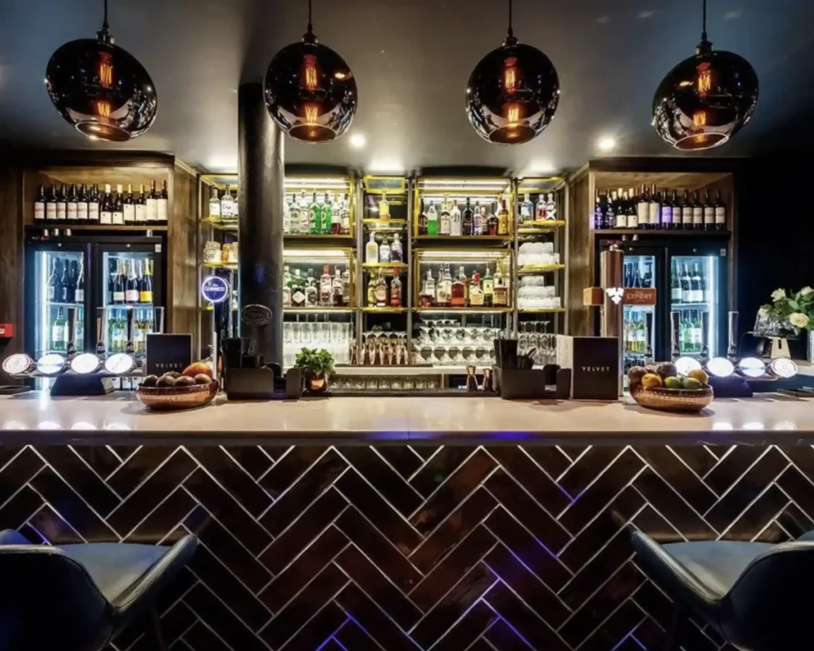 A stylish hotel bar with a sleek modern design, illuminated liquor shelves, and elegant pendant lighting.