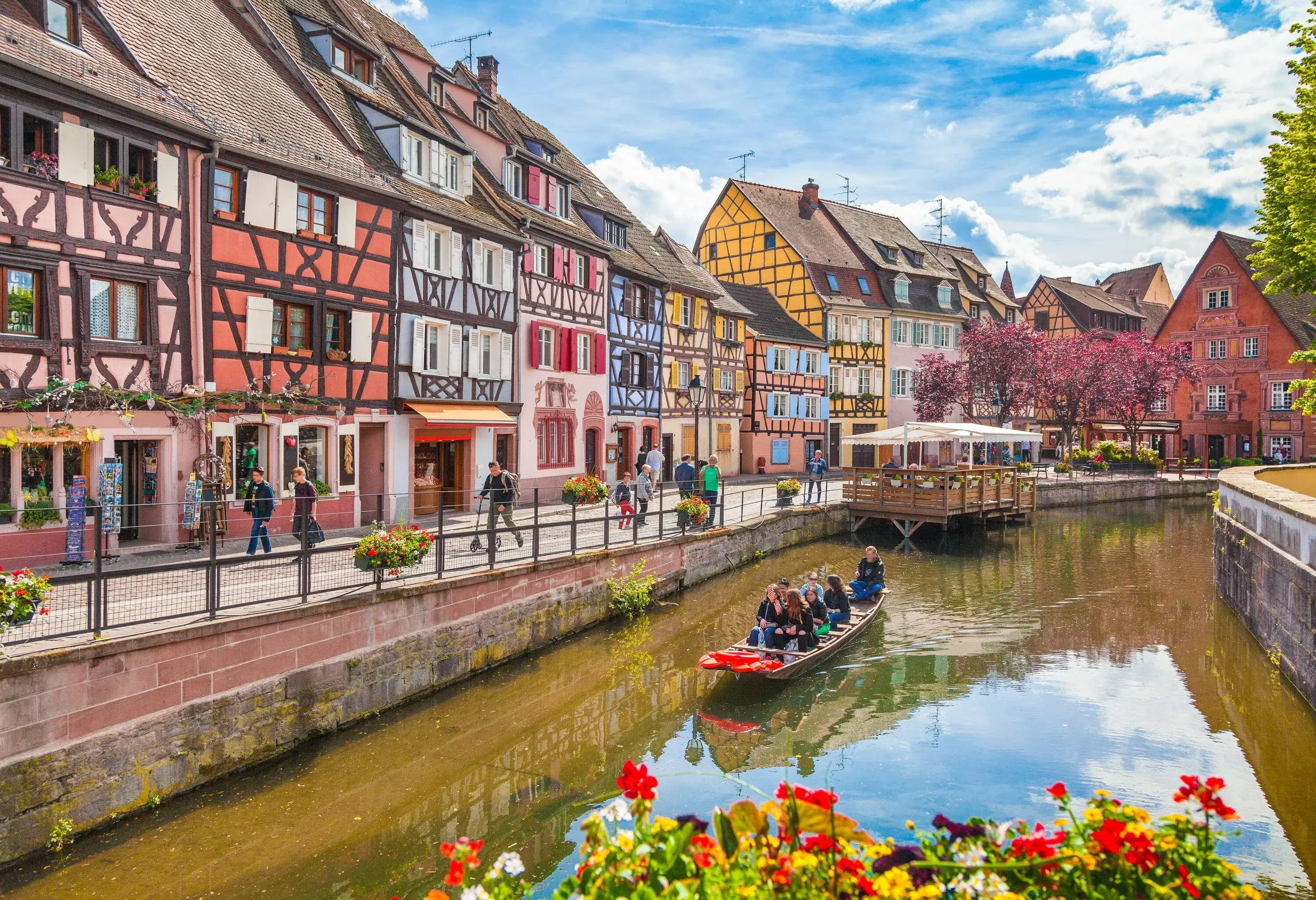 Tourists take a boat trip down a river that runs along a busy promenade lined with colourful half-timbered buildings.