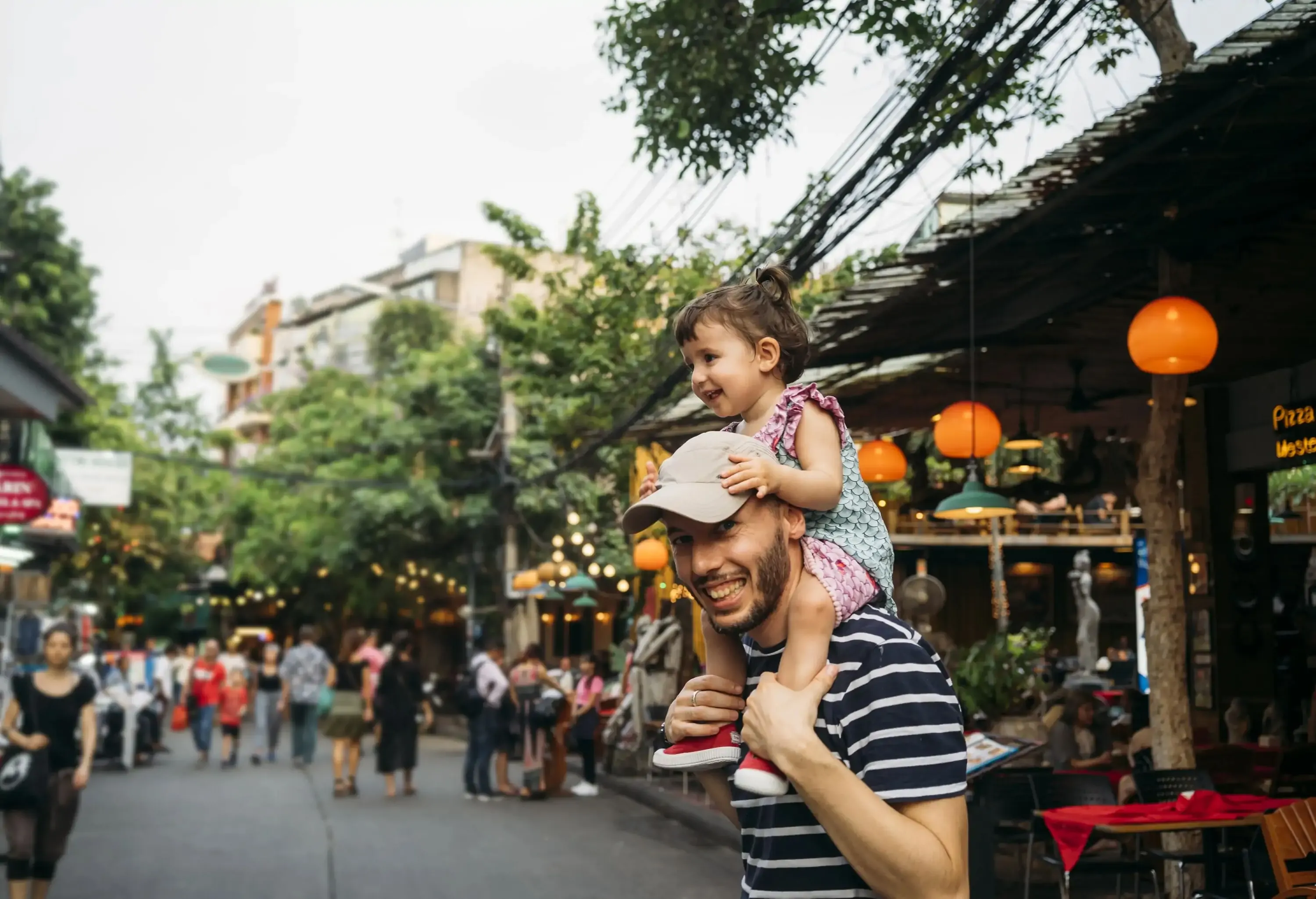 Smiling man in a striped shirt and cap stands on a street while carrying a toddler on his shoulders.