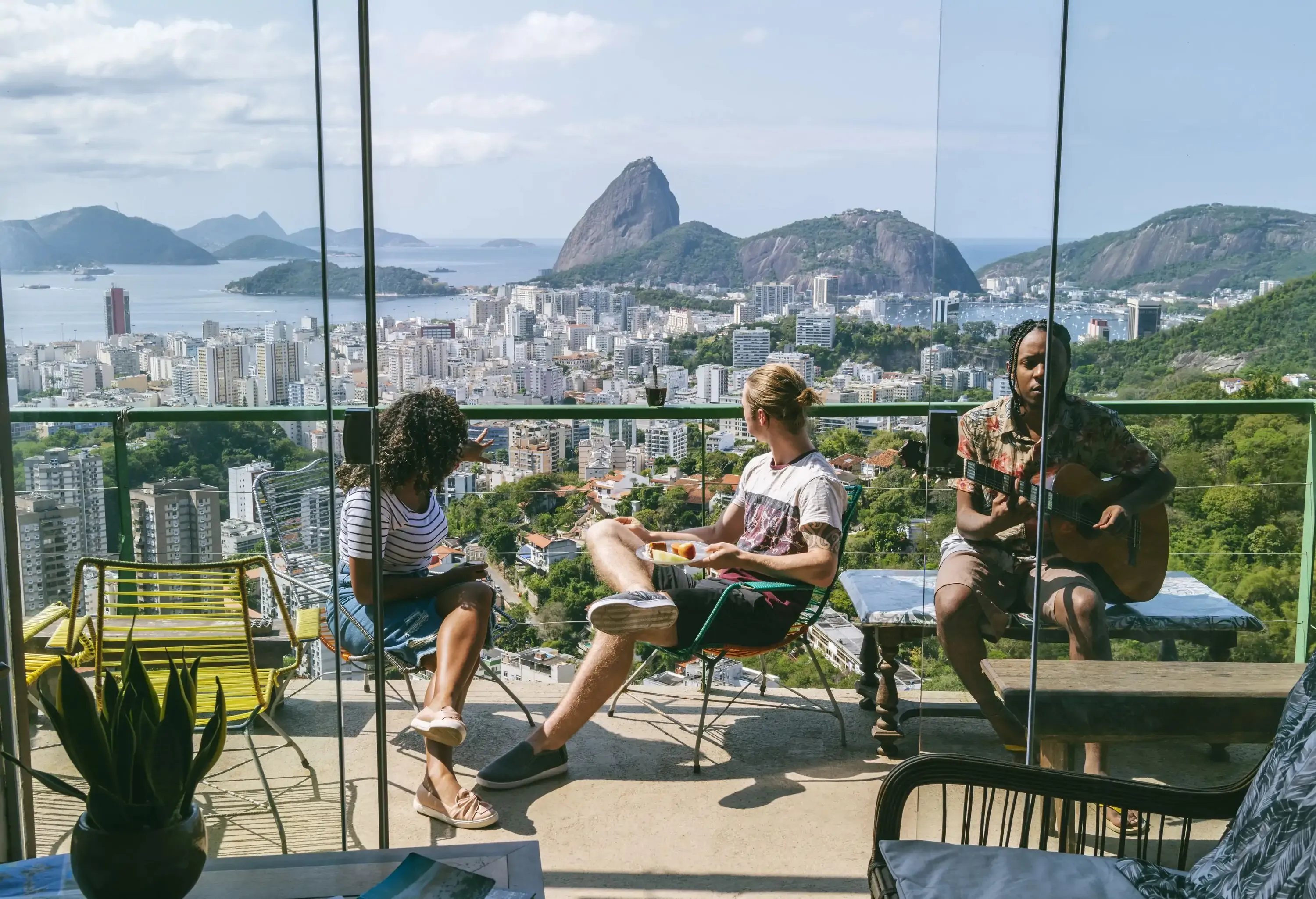 A diverse group of people on a balcony overlooking the beautiful coastal city with modern and towering buildings and rocky islands surrounded by the blue sea.
