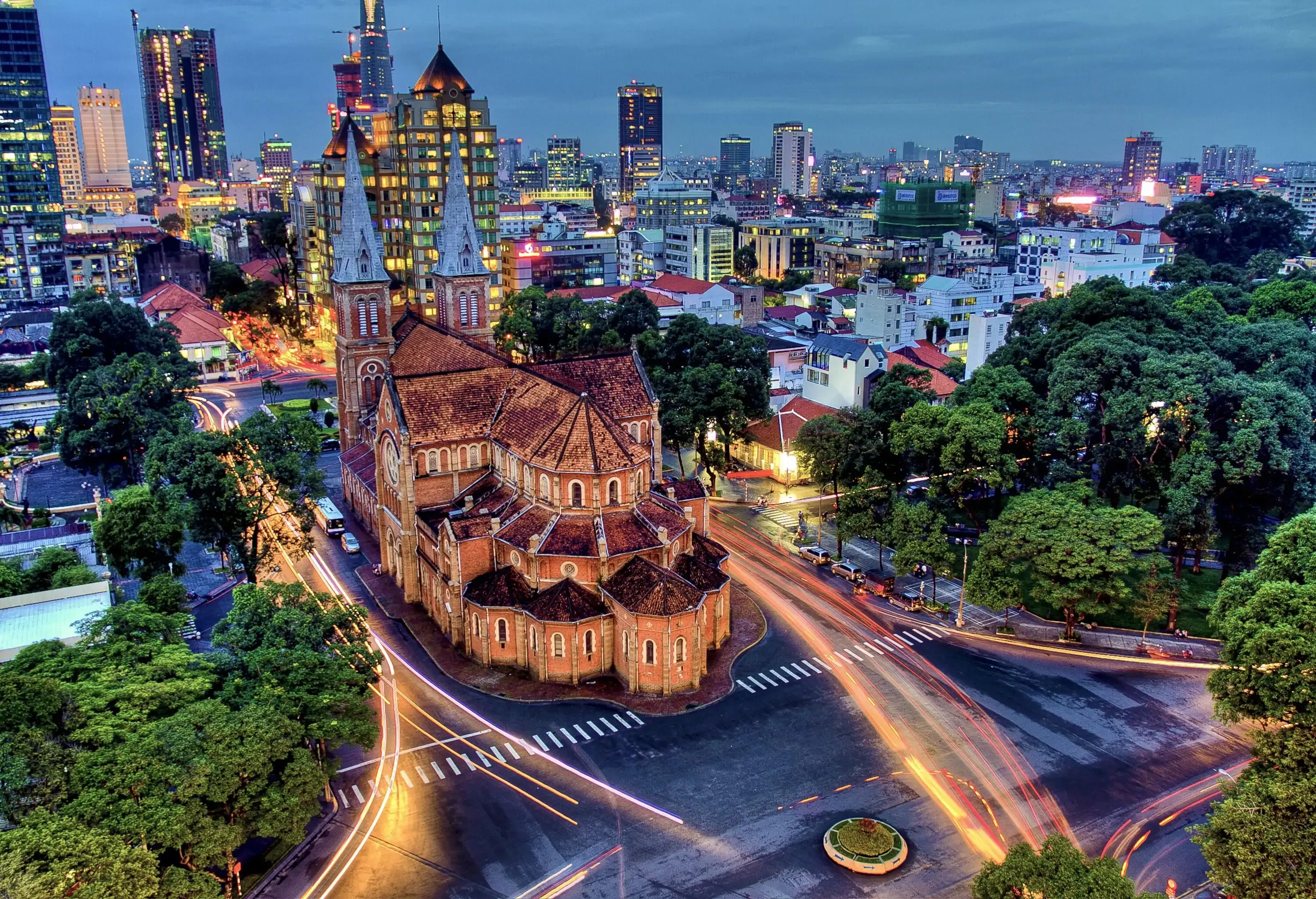 Notre-Dame Cathedral Basilica of Saigon, a red Romanesque brick cathedral with twin steeples in the middle of a busy intersection, forested parks, and bright city buildings.