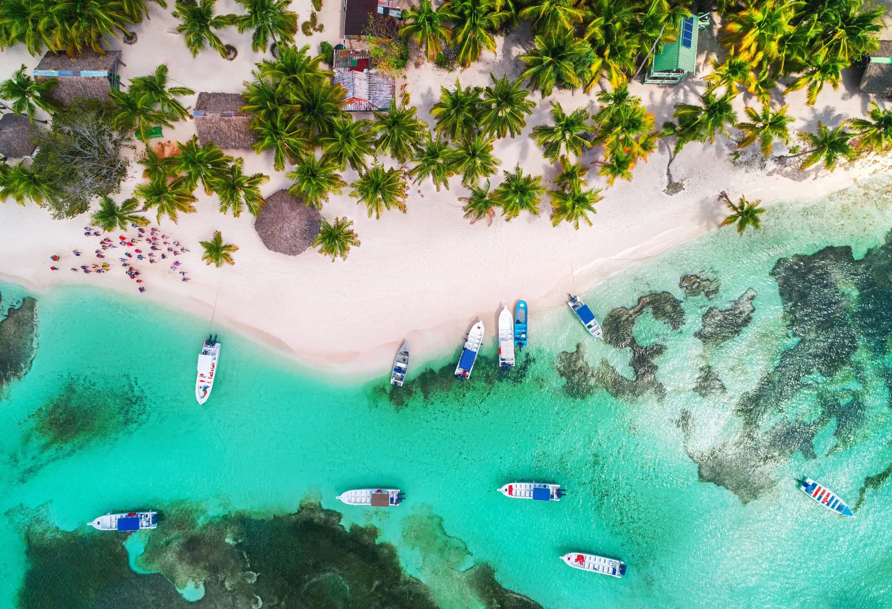 A white sand beach with multiple boats on turquoise waters and cottages under a grove of palm trees.