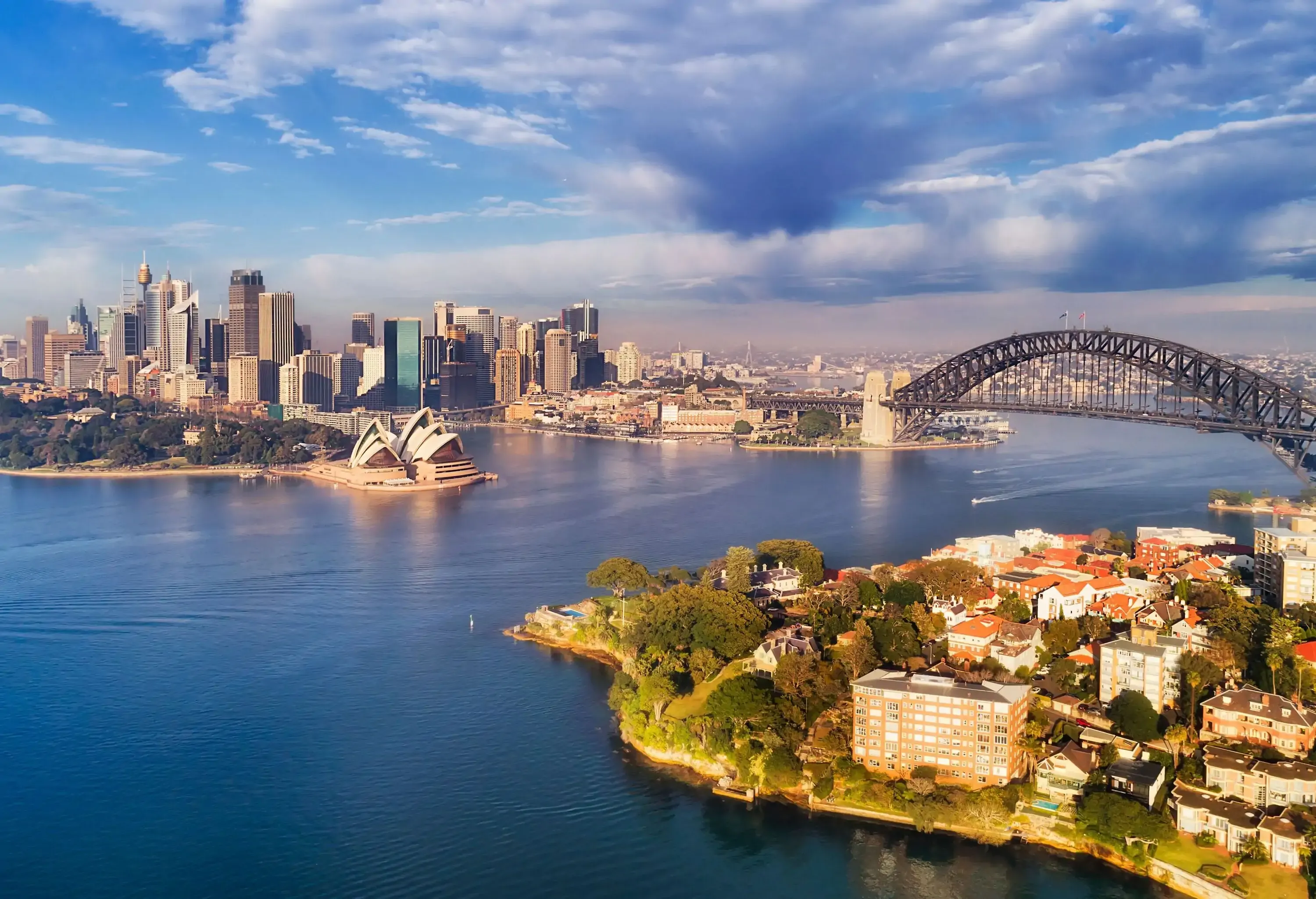 A steel arch bridge spanning the harbour near the iconic Opera House on the foreshore against the city skyline on a dramatic sky.
