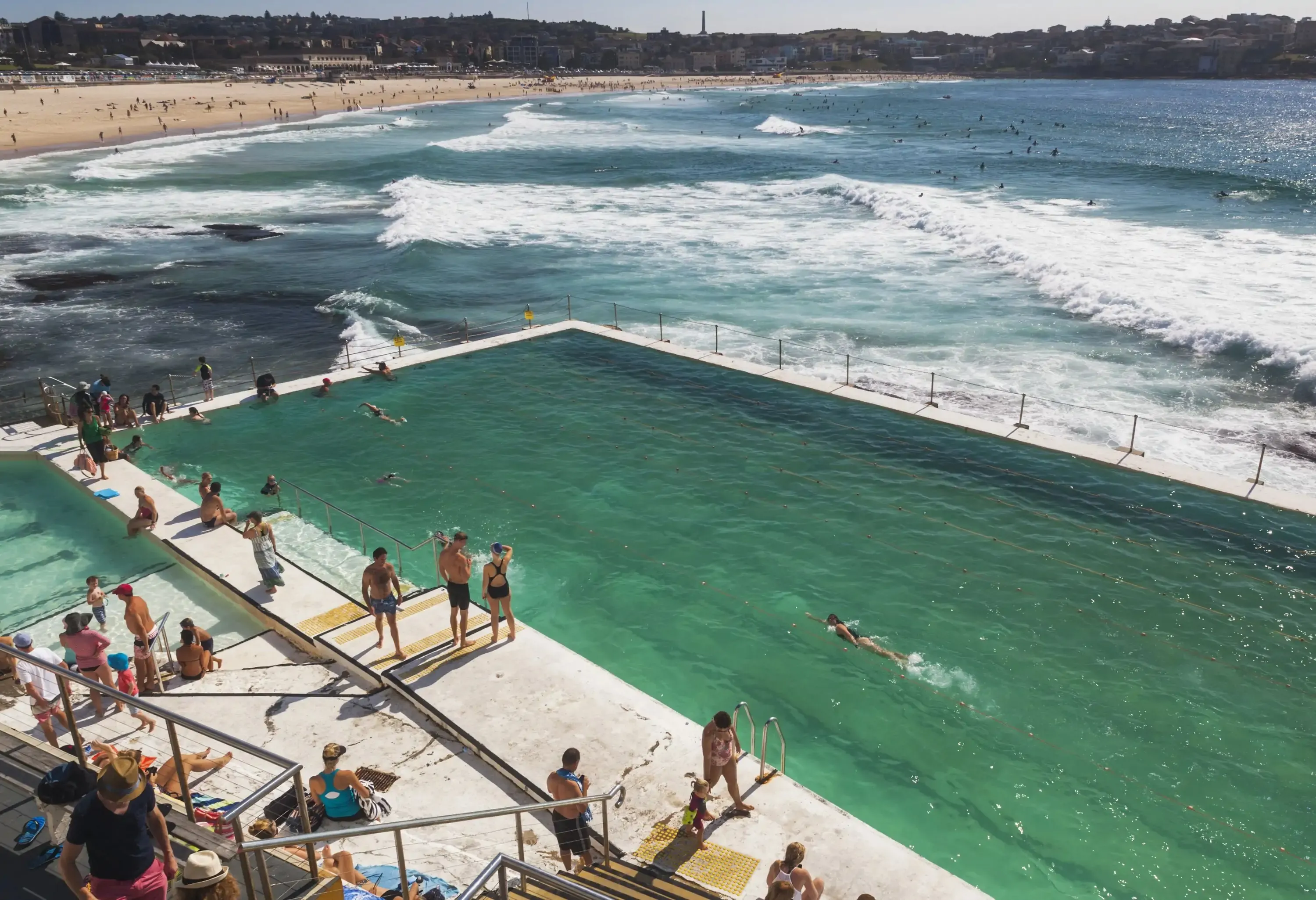 Many people are on the beach enclosed by mountains and in the pool next to the sea with the rolling white waves.