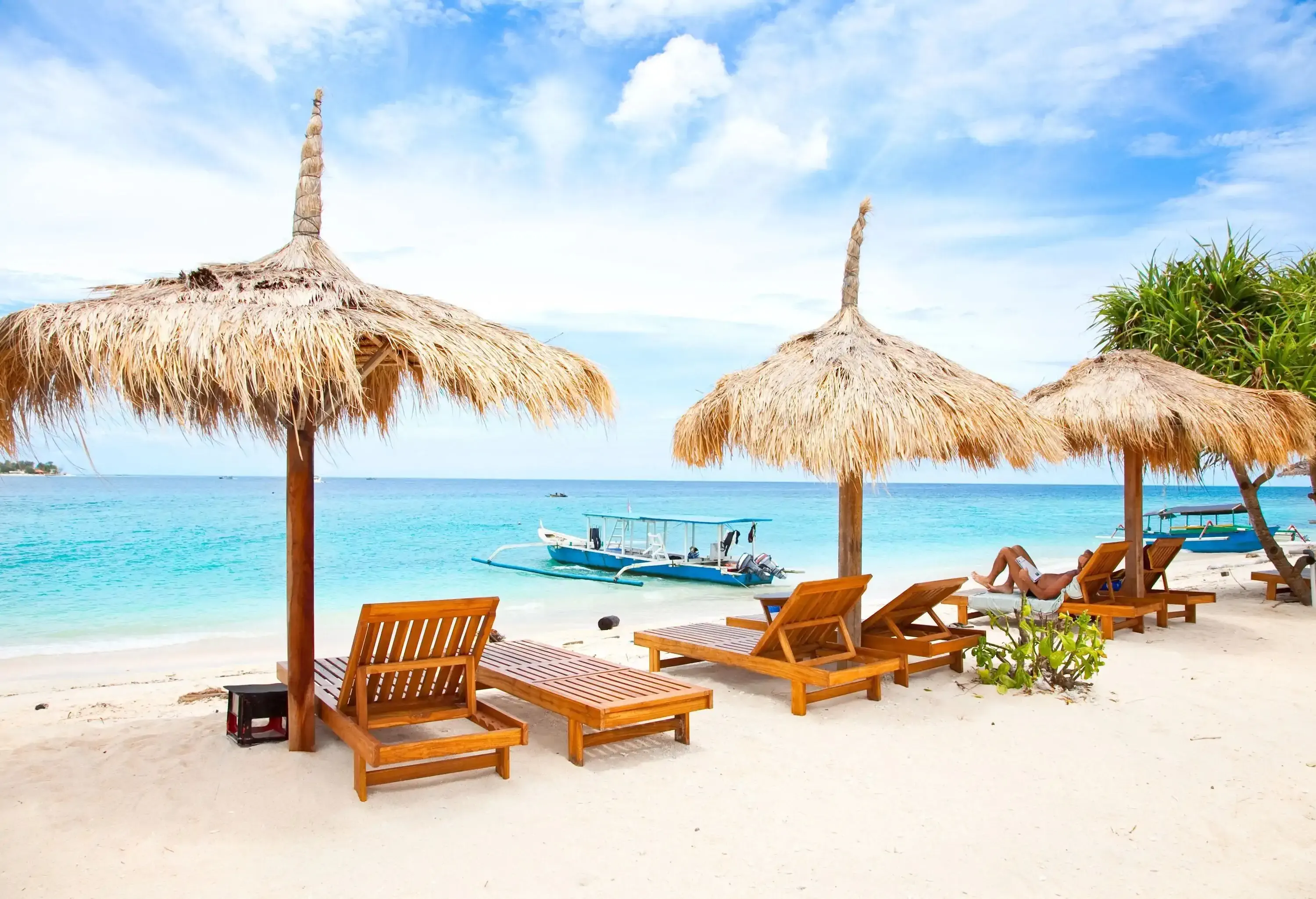 A pristine beach with inviting thatched parasols and comfortable wooden sun beds, where a sleek double outrigger boat floats serenely on the crystal-clear waters beyond.