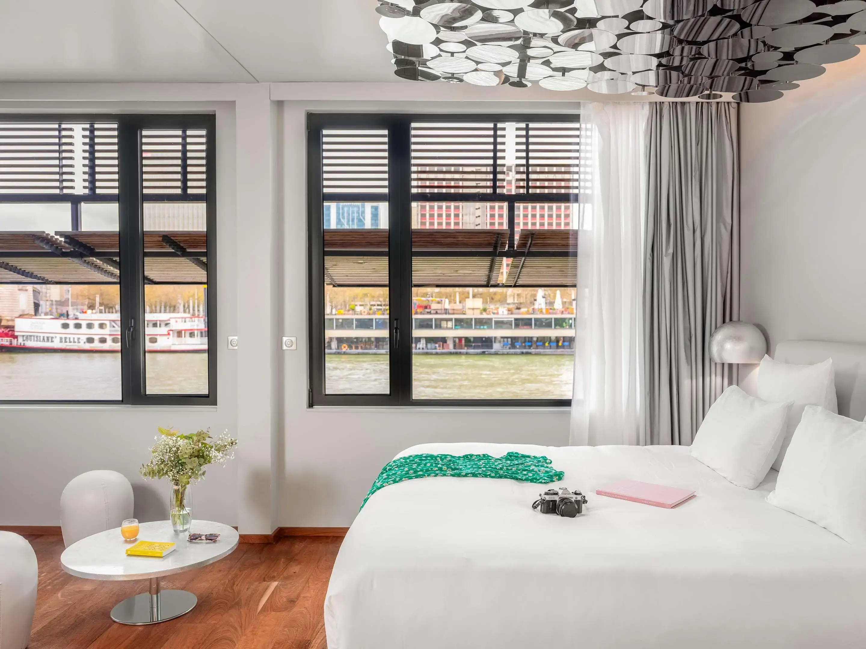 A modern hotel room with a large bed, a small table with flowers and a drink, windows overlooking a river and boats, and city buildings visible in the background.