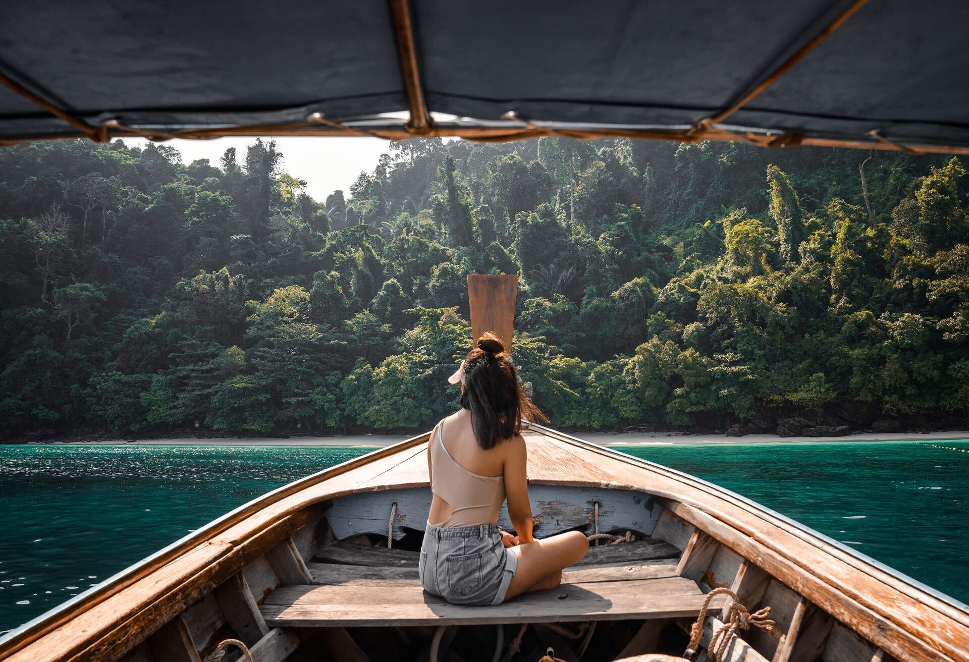 A young lady sits on the stern of the boat approaching the forested shore.