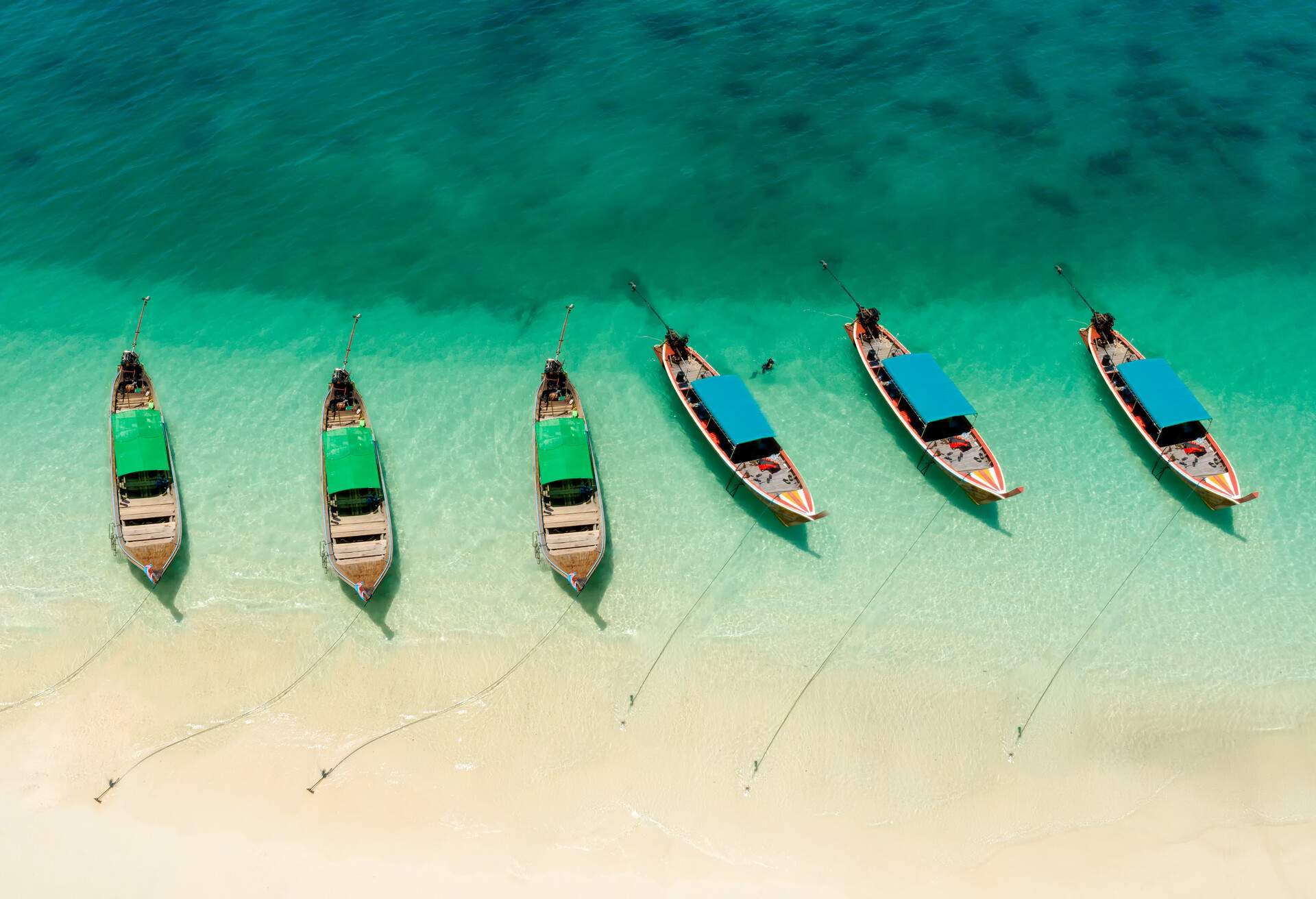 Six long-tail boats with red and blue roofs anchored on a pristine beach.