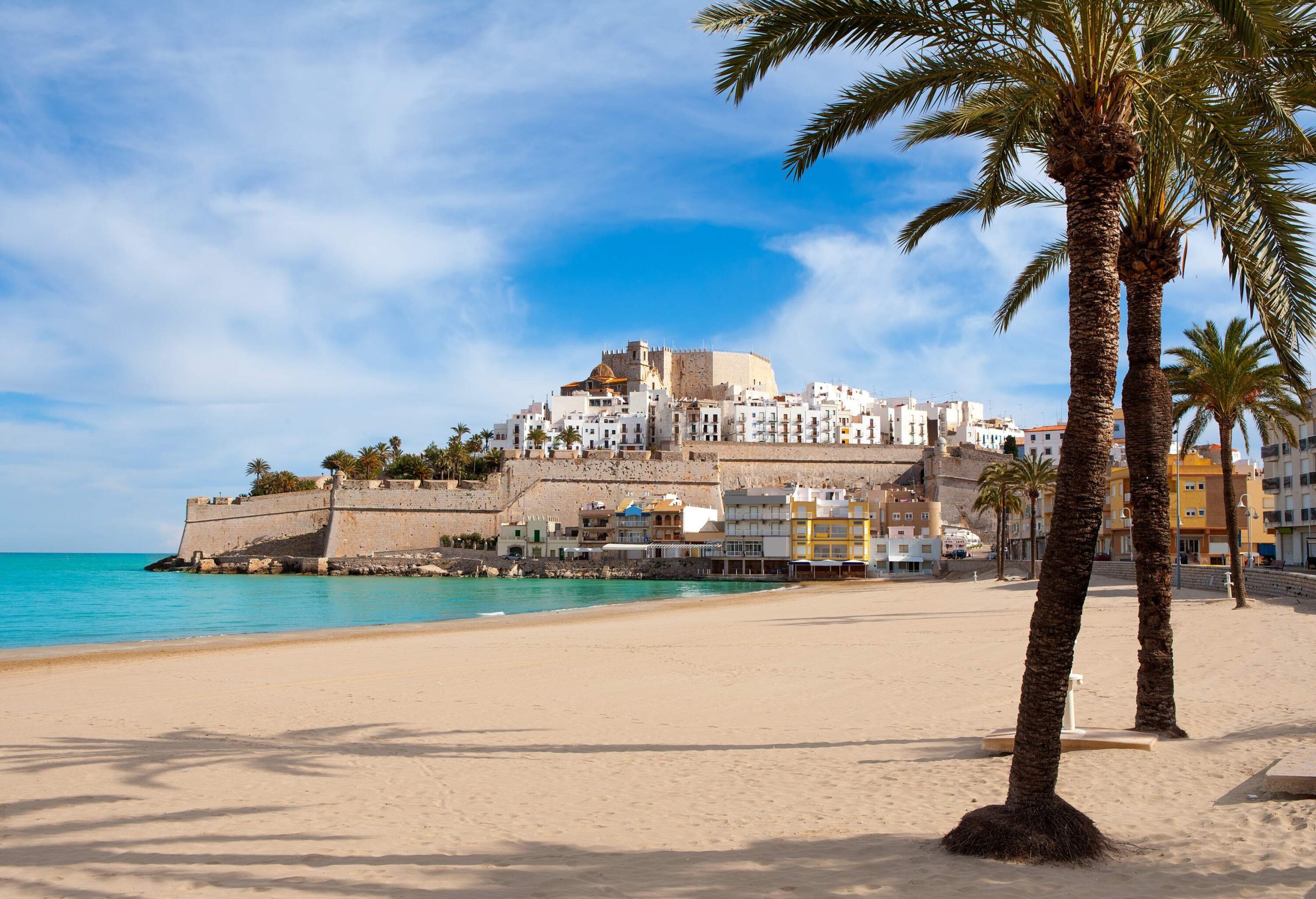 A castle surrounded by white houses on top of a crag overlooking a white sand beach with calm turquoise water.