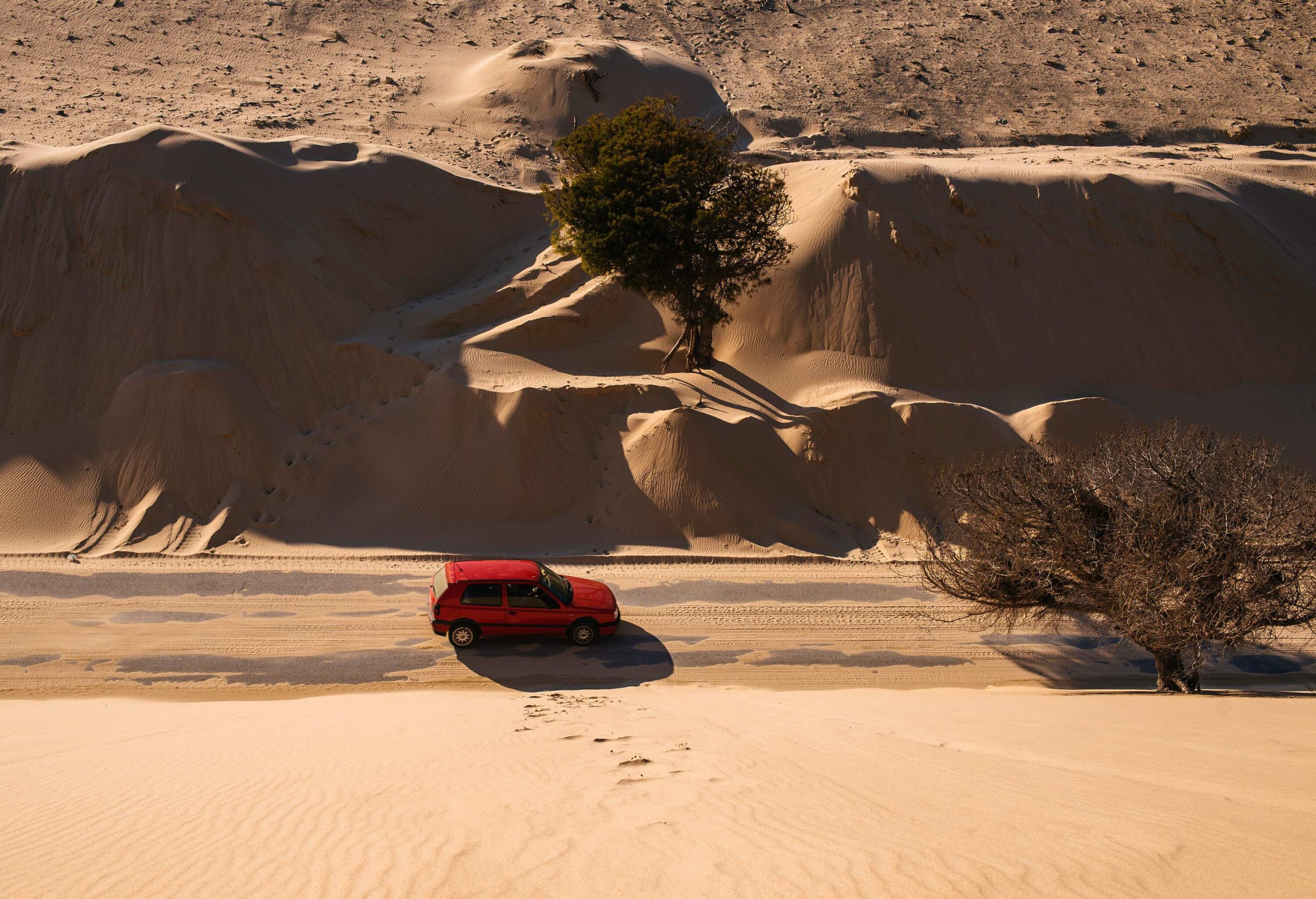 A red car travels on a concrete road with sand through the dunes.