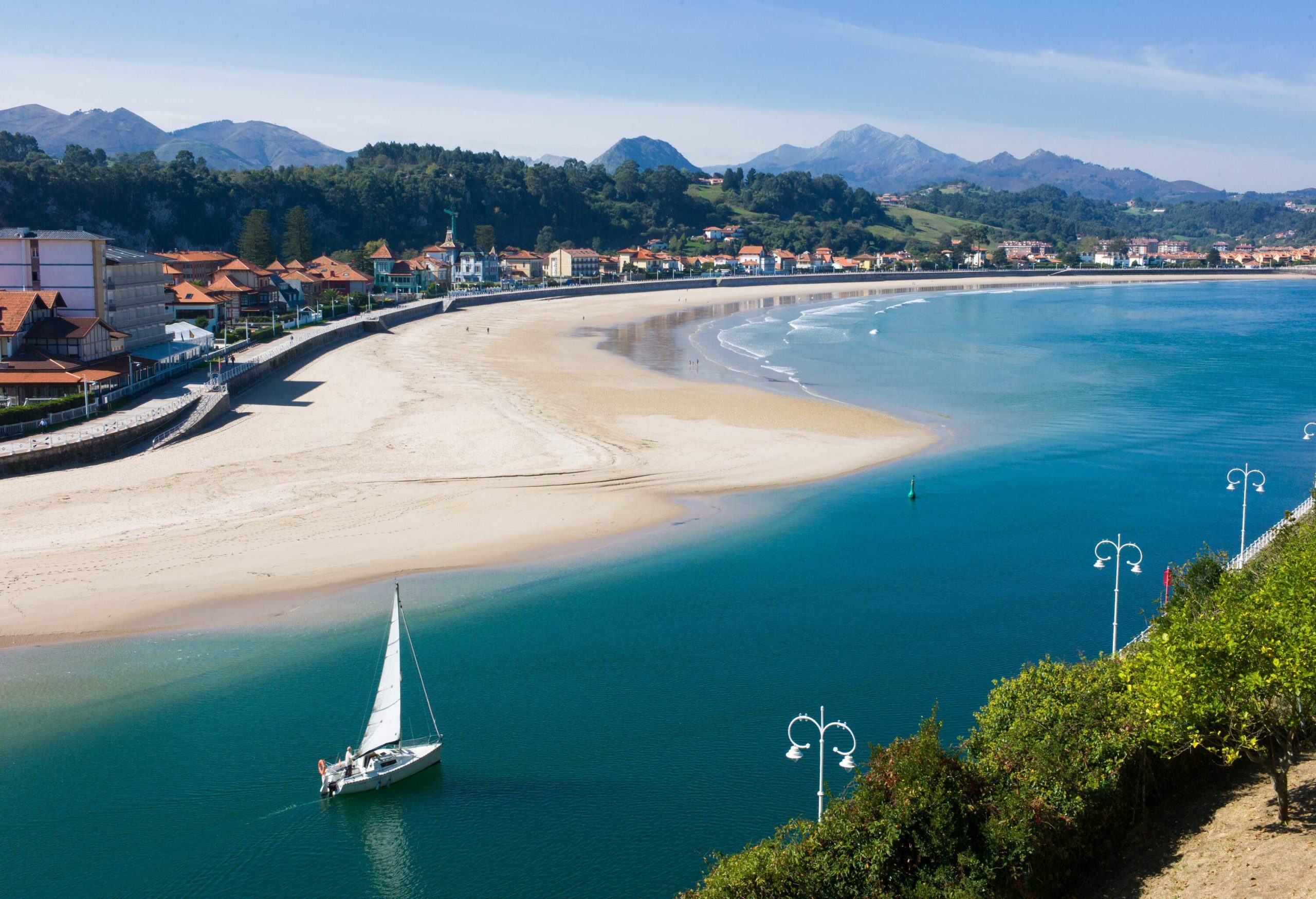 A sailboat cruising along a white sand beach with waterfront buildings.