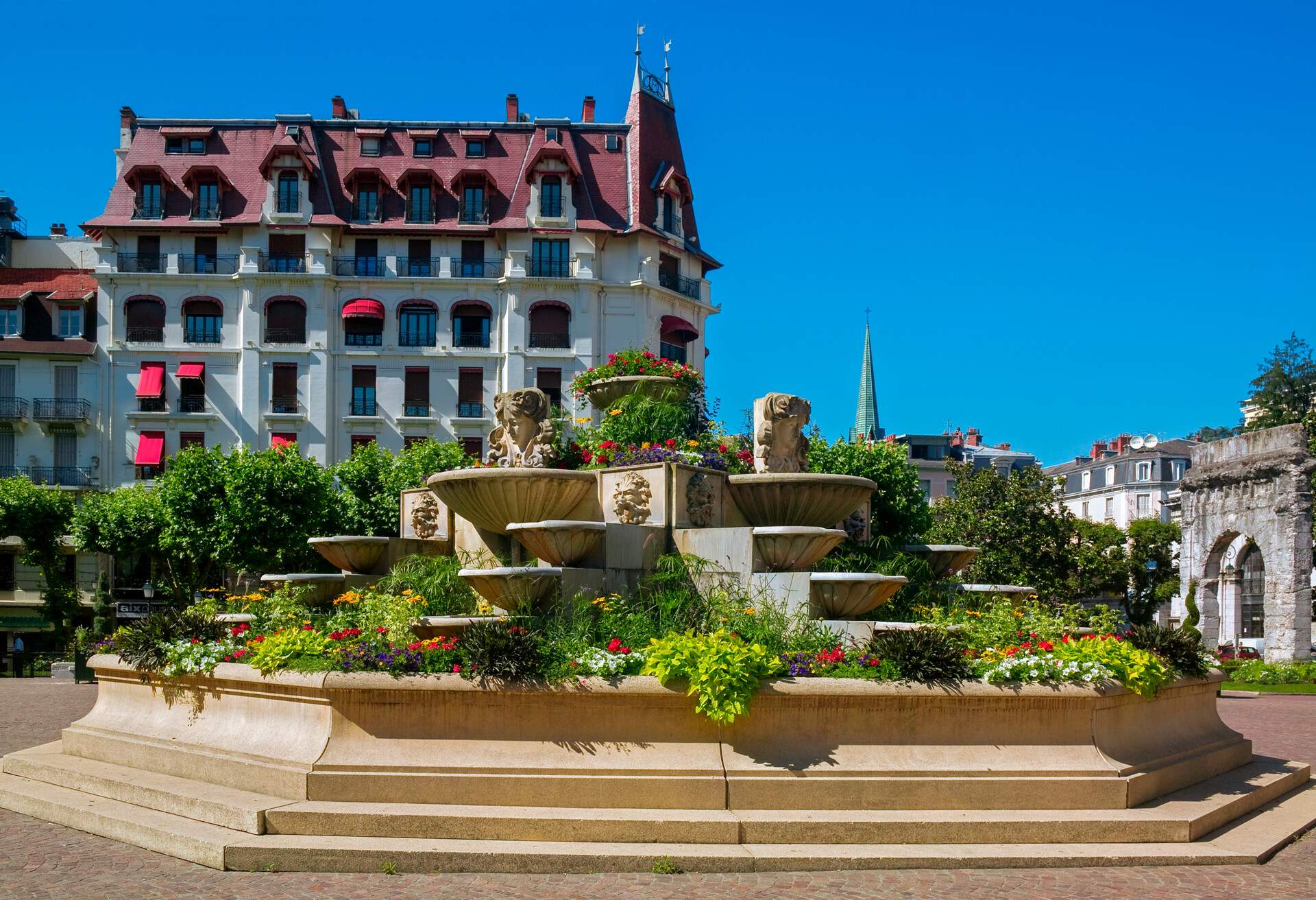 A multi-story white mansion in front of a plant-decorated fountain beside an antique Roman arch.