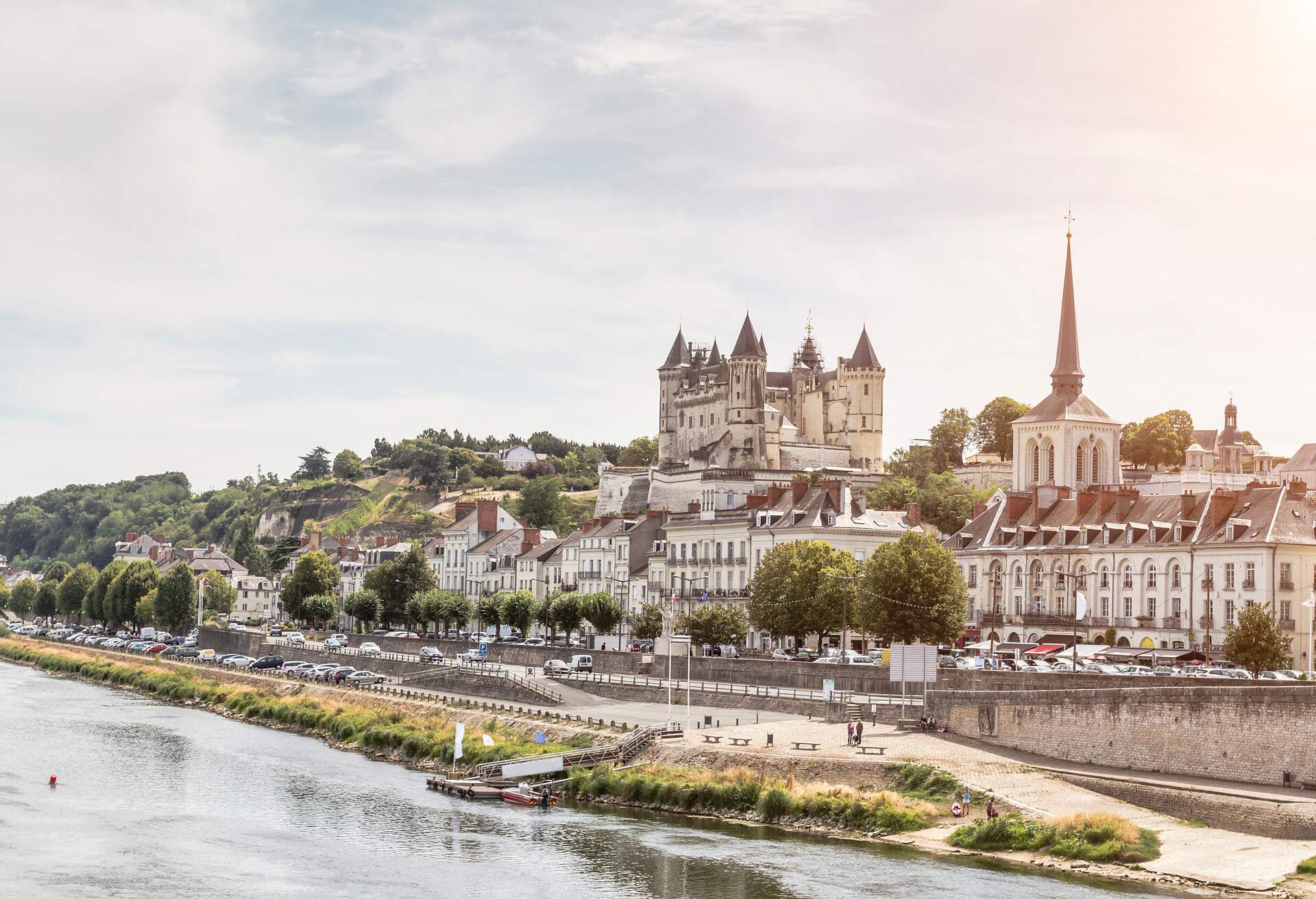 An old riverside city with classic buildings and a fortified medieval chateau on top of a hill against the pinkish-bluish sky.