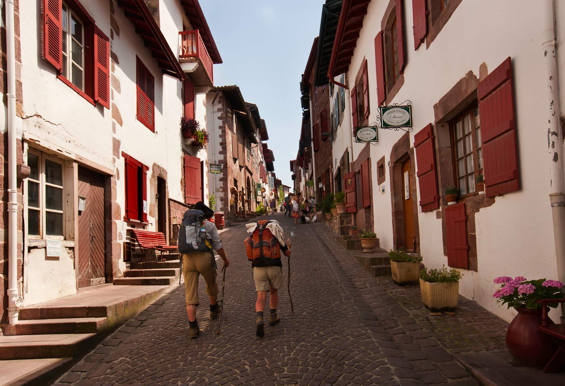 Two people with rucksacks and trekking poles walk along cobbled streets lined with old traditional houses with red windows.