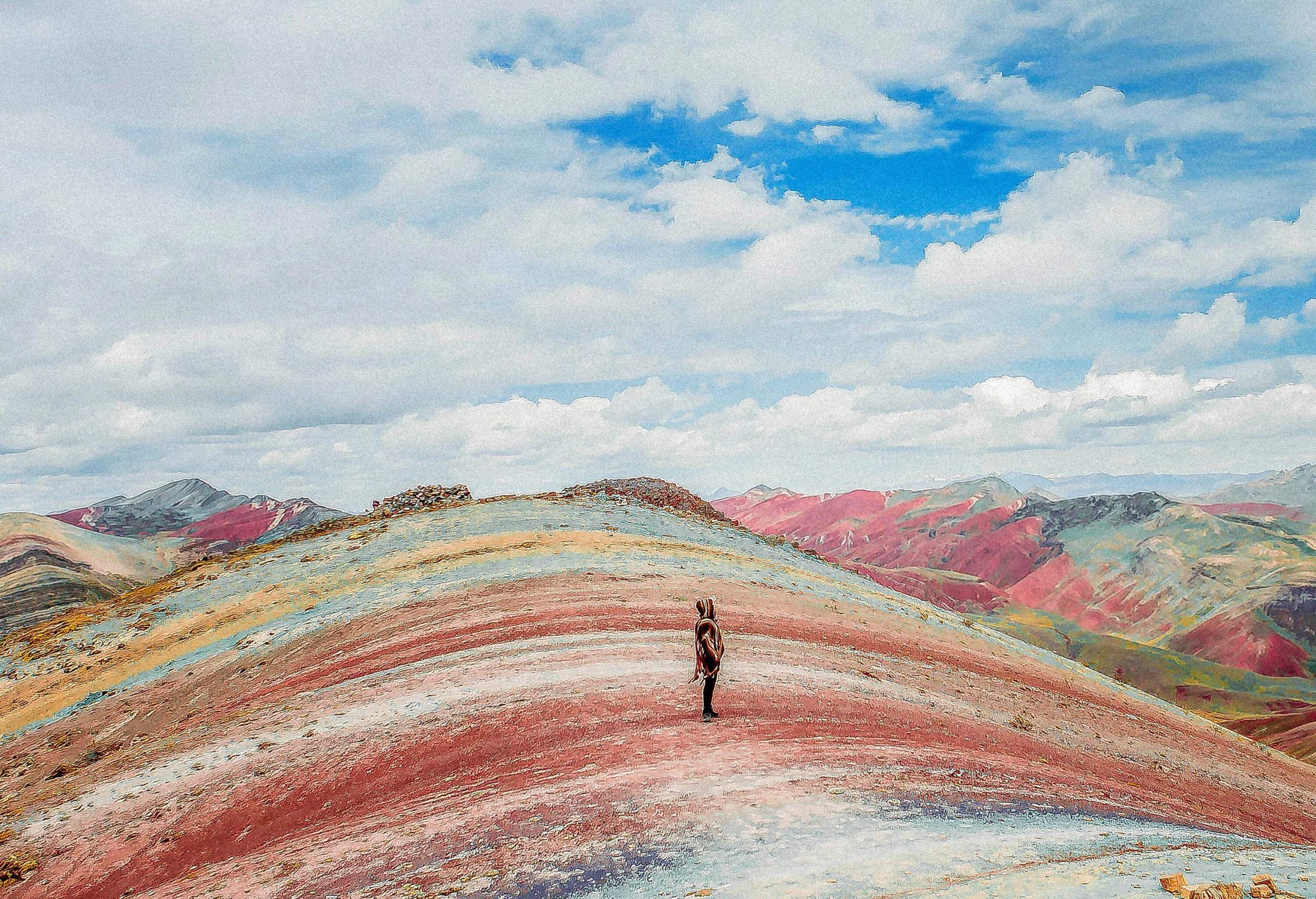 A tourist standing atop a spectacular rainbow-striped hill against the white cloudy sky.