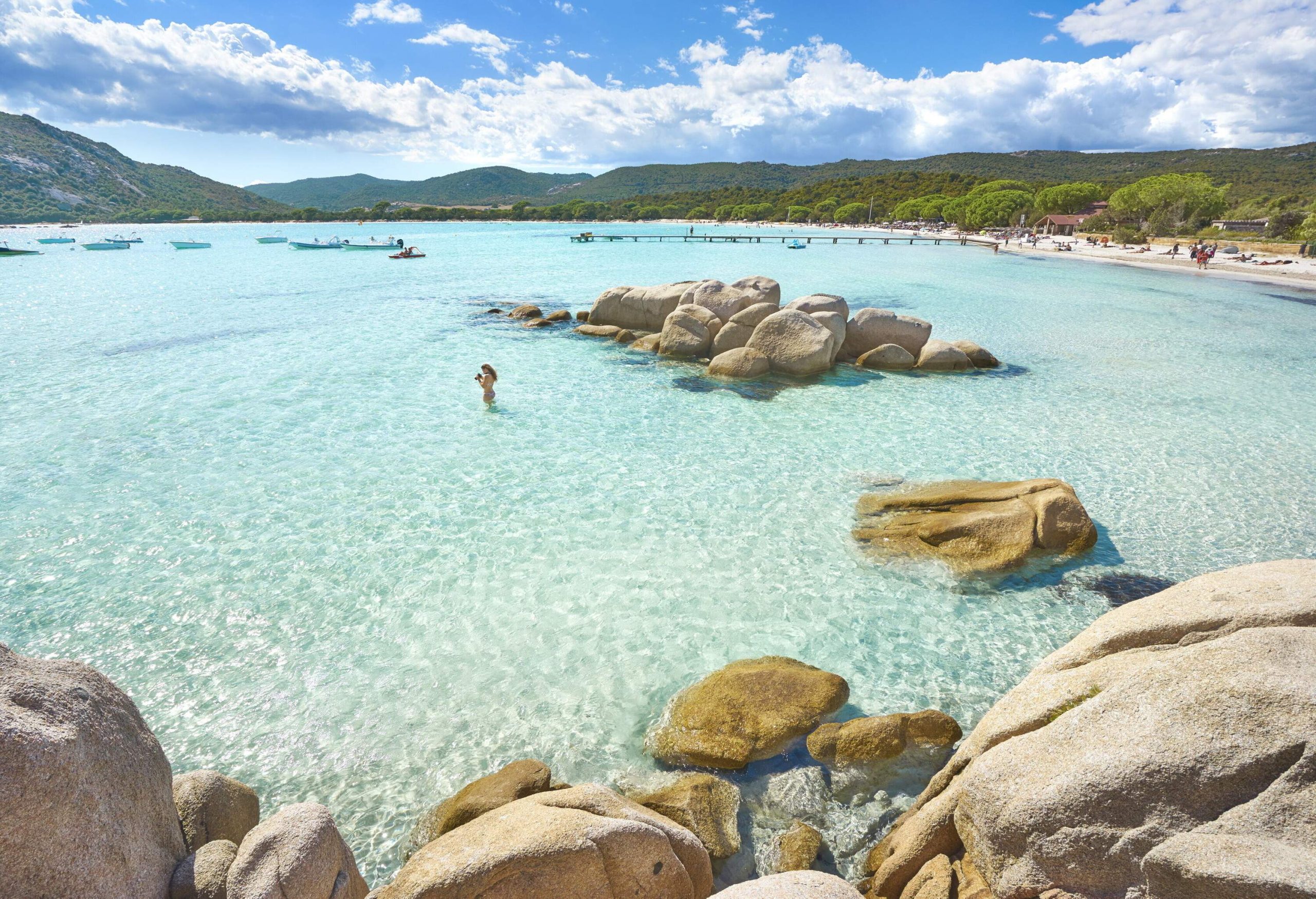 A person stands in crystal-clear water as seen from a rocky coast.