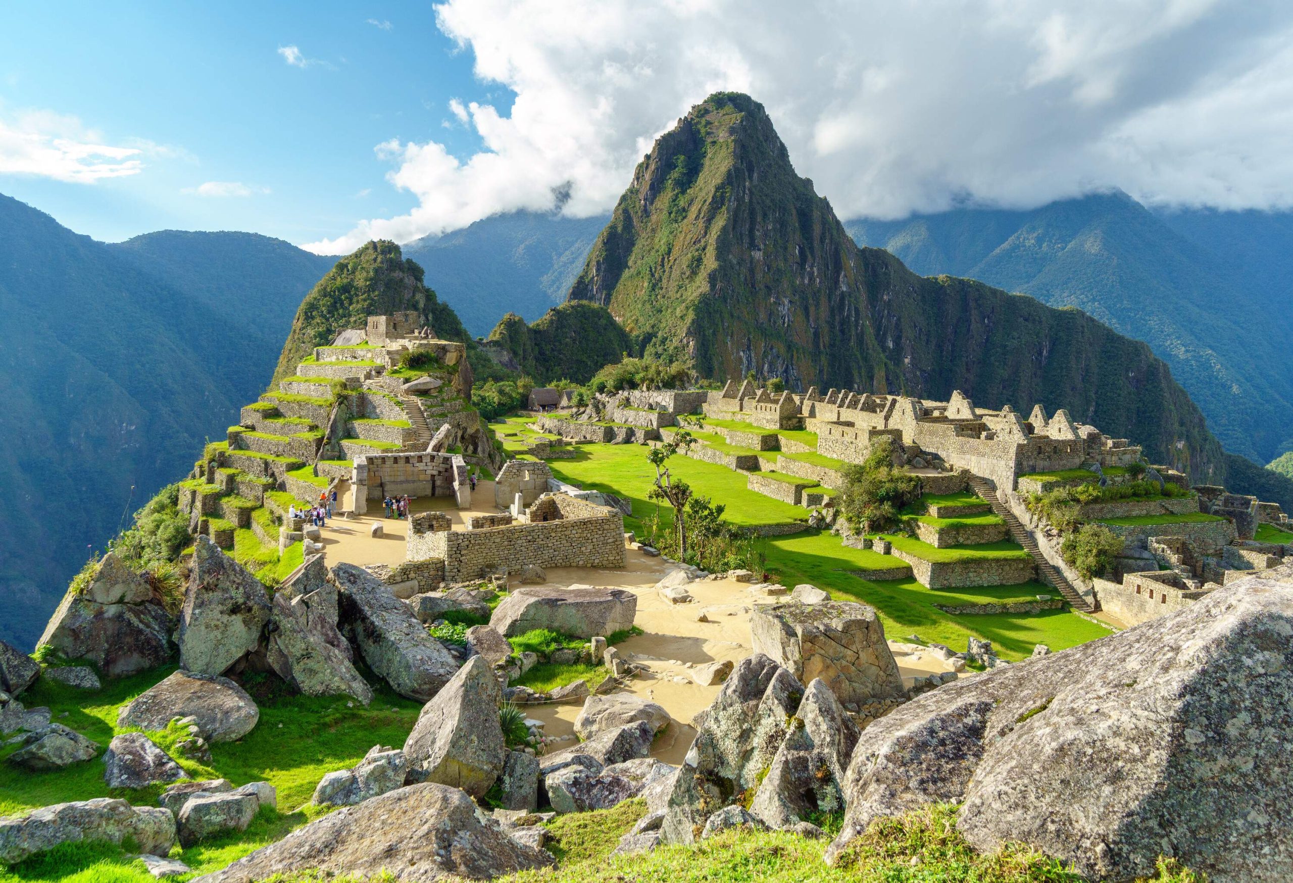 Boulders and rocks in the foreground offer a rugged perspective as the iconic Machu Picchu emerges on a vibrant green landscape, embraced by majestic mountains in the backdrop.