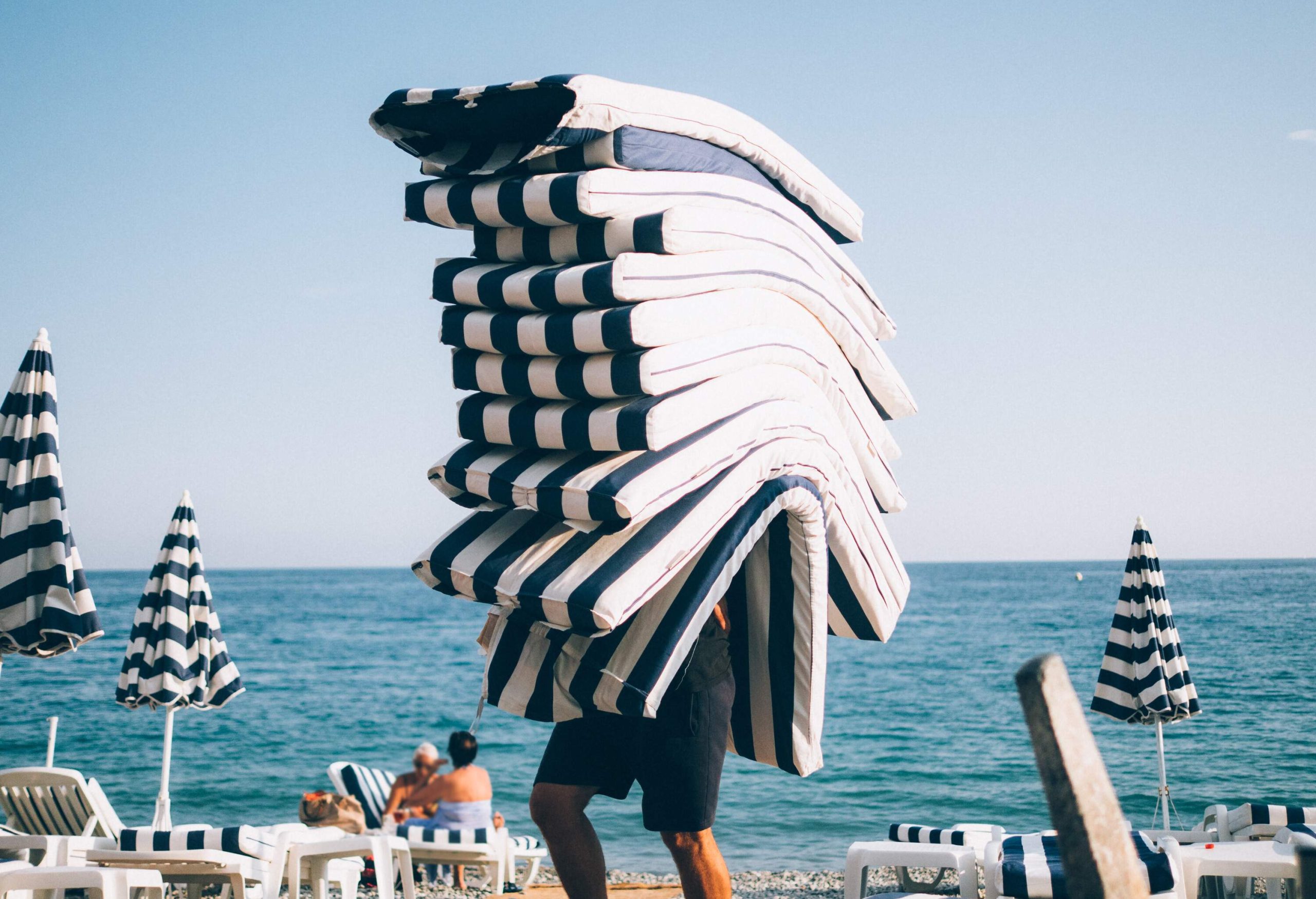 A man carrying a pile of blue and white striped sunbed cushions across the beach.