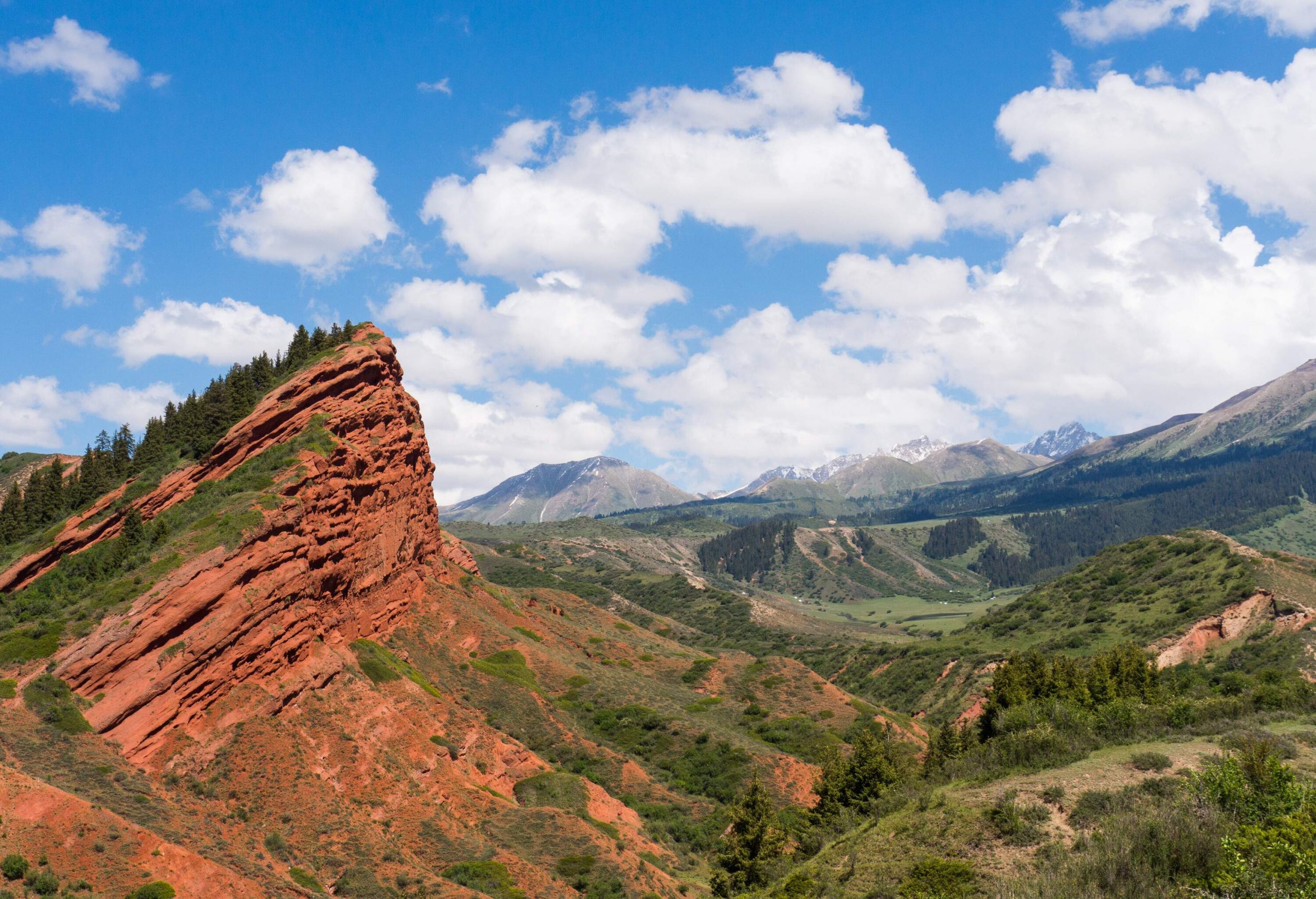 A rock mountain with jagged peaks overlooks a mountainous valley covered in lush trees.