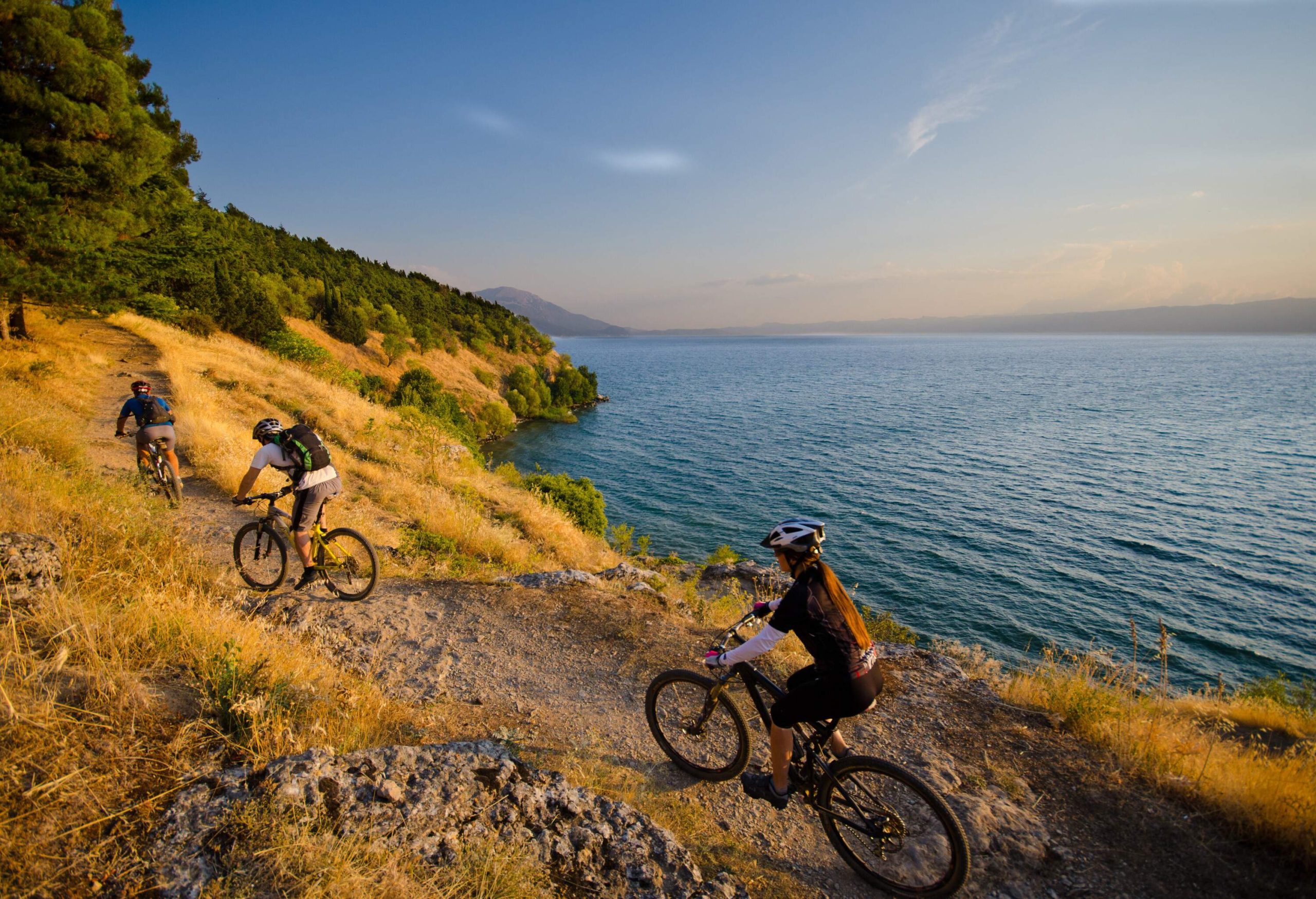 Three cyclists ride on dirt roads in lush hills by a tranquil lake at sunset.