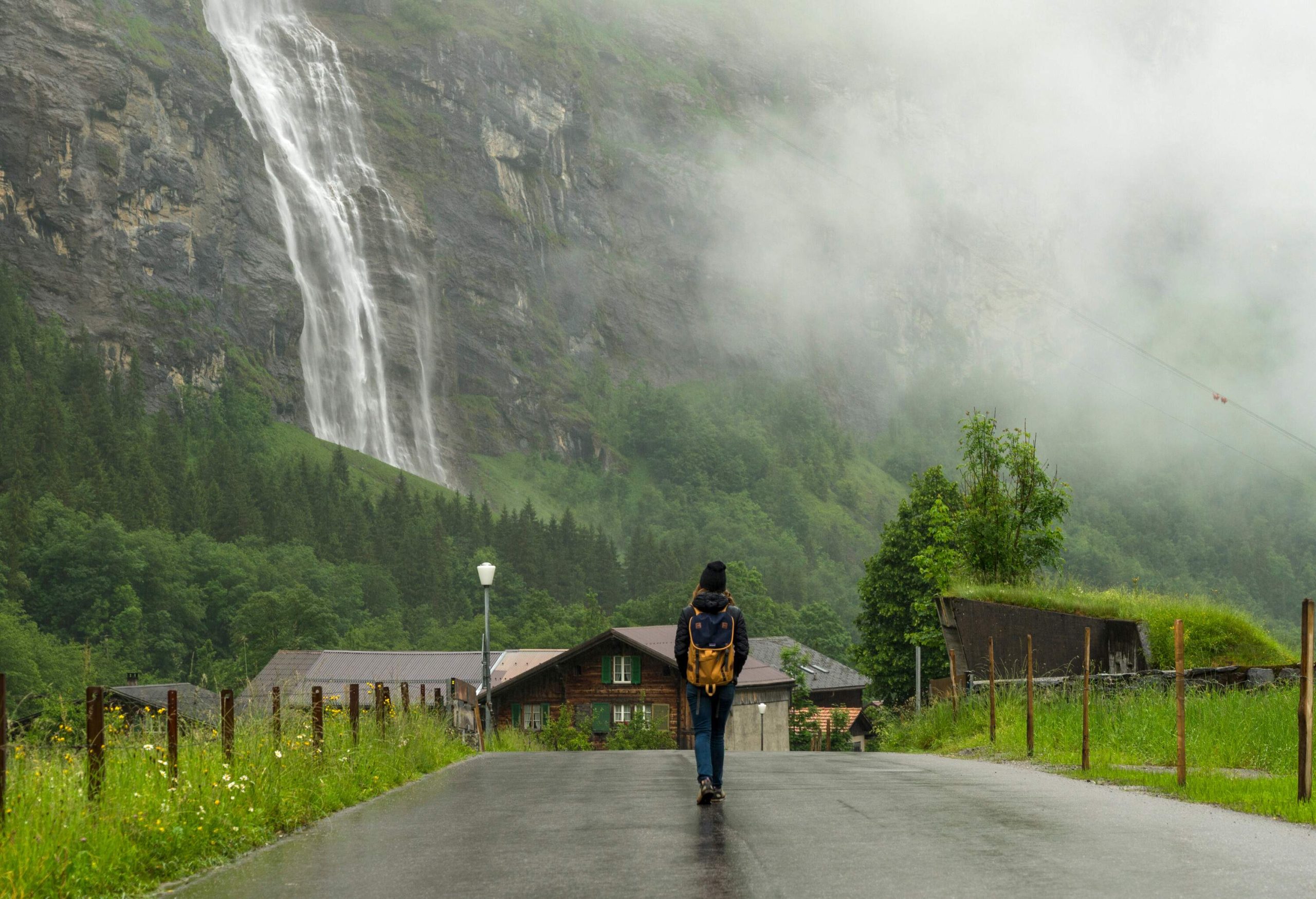 A person walking on a wet road toward the houses along a misty valley with waterfall gushing down a steep rock face in the distance.