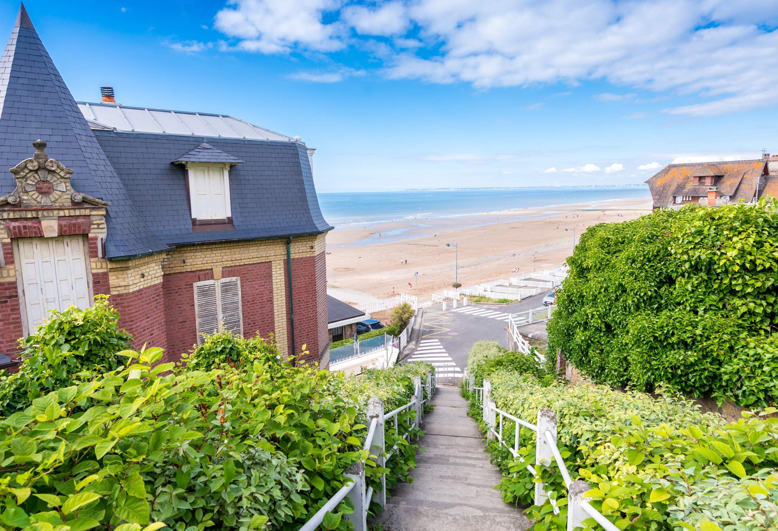 A downhill stairs bordered with lush plants and brick houses in a neighbourhood that leads to a roadside beach.