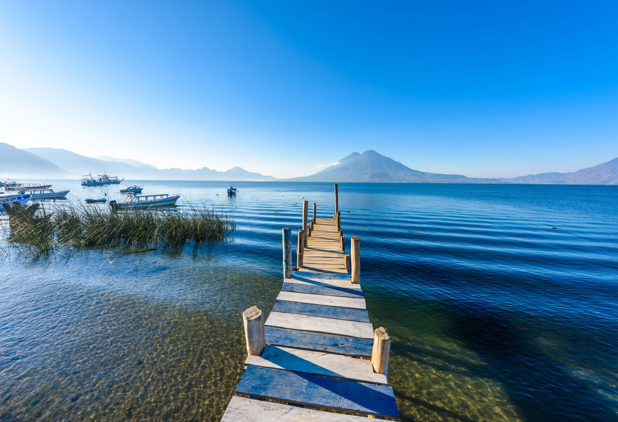 A wooden walkway runs directly over the clear lake, where boats are moored and a cone-shaped volcano may be seen in the distance.
