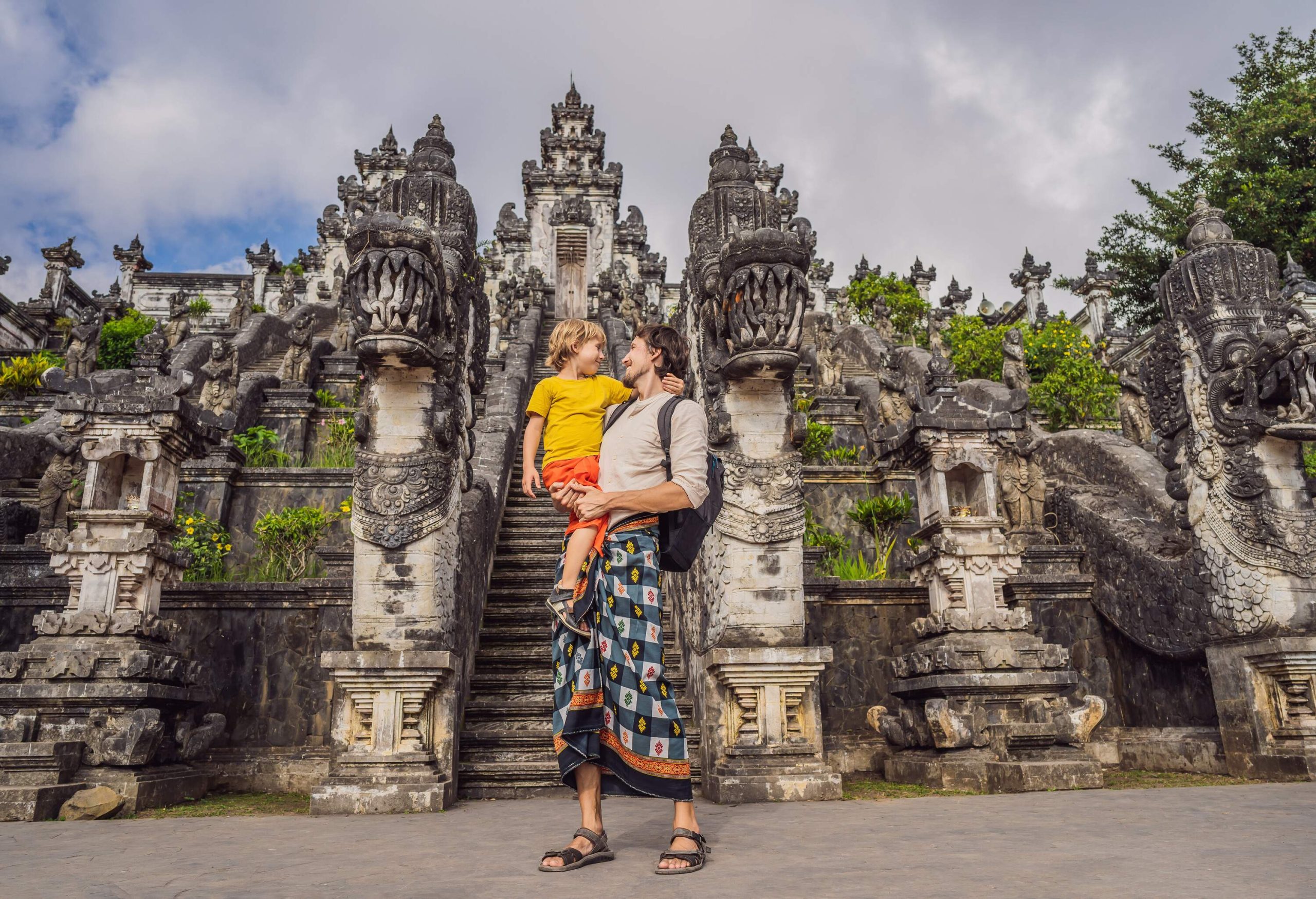 A man carrying a little boy with a background of three dragon stairs leading to the temple.