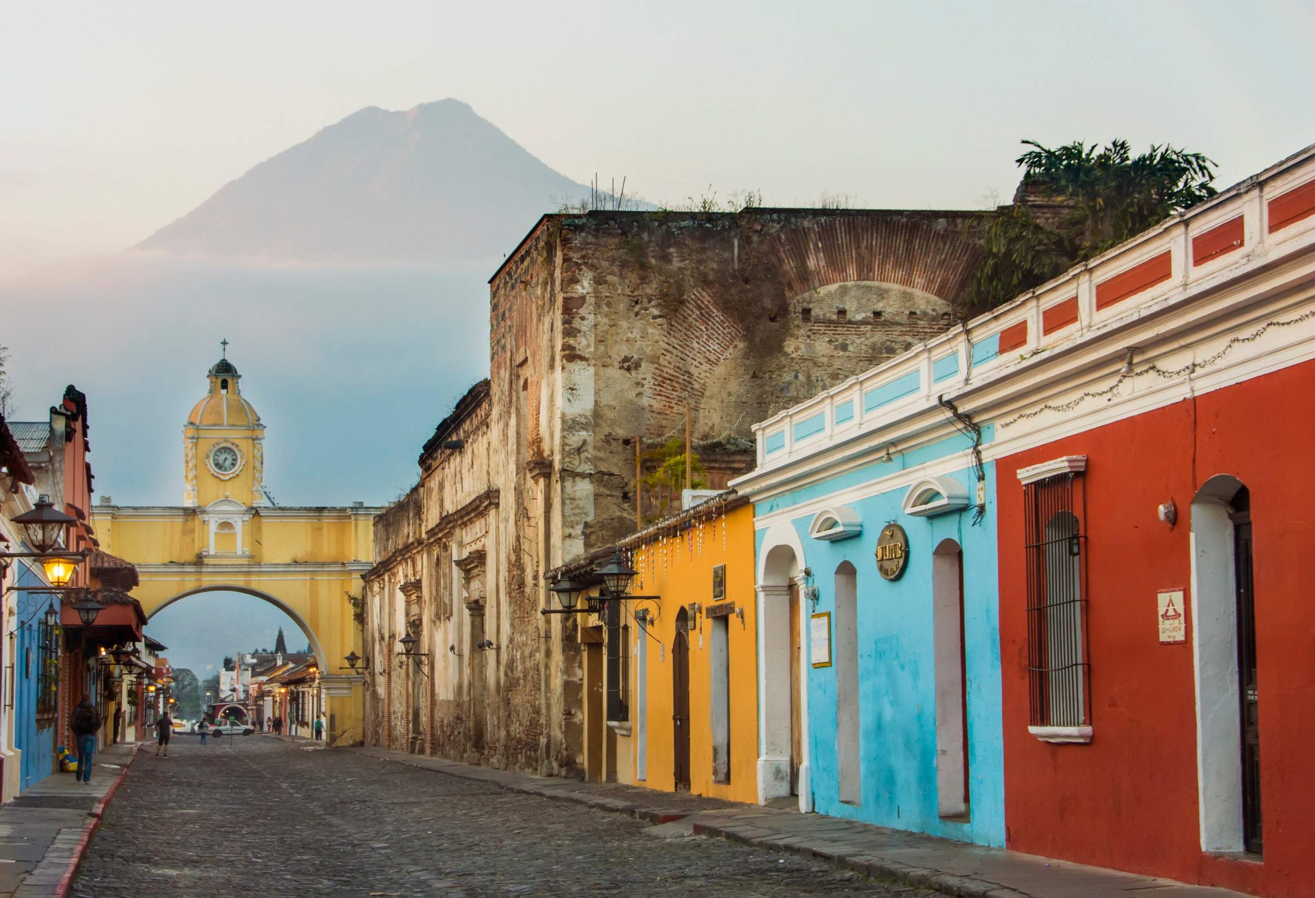 A charming cobblestone street meanders through a row of vividly painted houses with flat roofs, guiding the way towards a vibrant archway where a mist-shrouded volcano looms mysteriously in the distant backdrop.