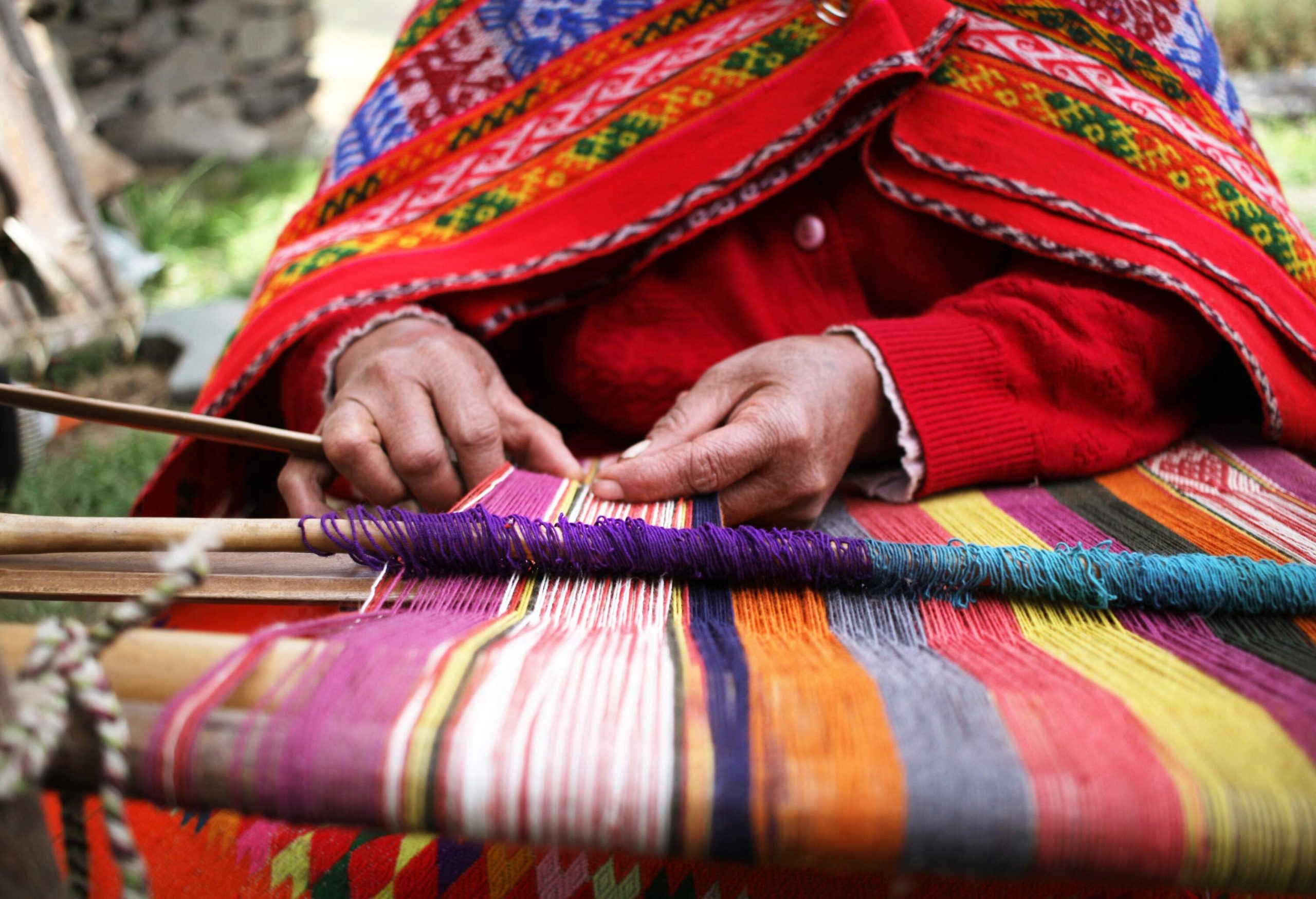 A person wearing a colourful shawl and jacket weaves the fibres of colourful fabrics into a traditional weaving machine.