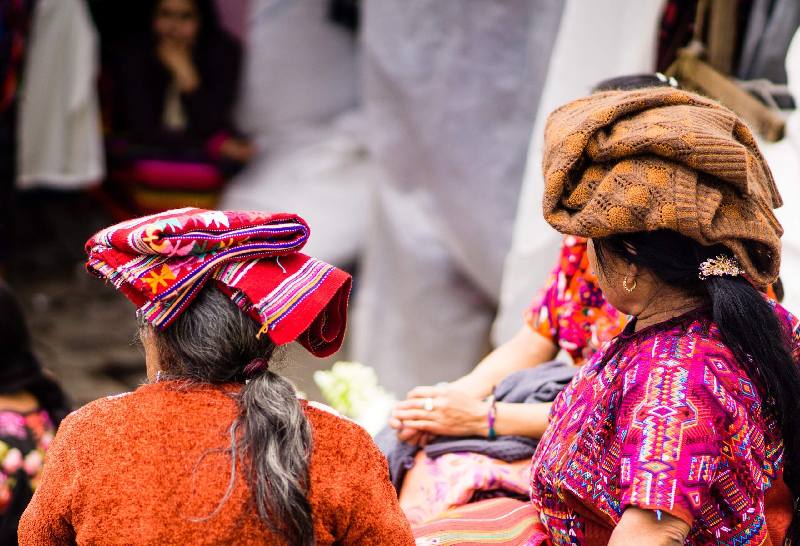A group of women, their heads adorned with vivid, colourful cloths, browse a bustling market as they gather in front of a vibrant cloth shop.