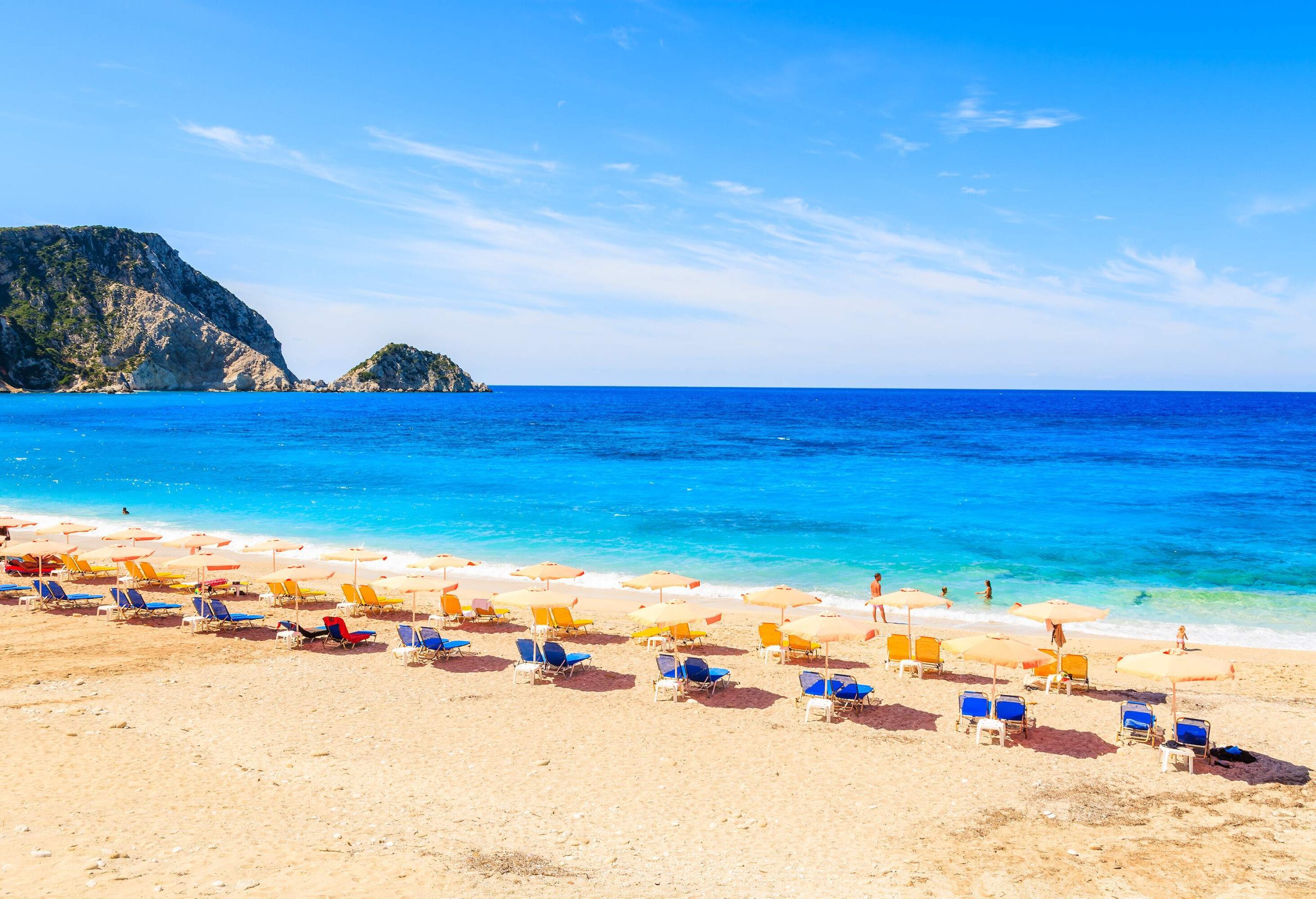 A stunning beach with rows of colourful parasols and blue sun loungers, where beachgoers relax and enjoy the soft waves and crystal-clear blue sea, is set against the backdrop of a steep and rugged rock formation.