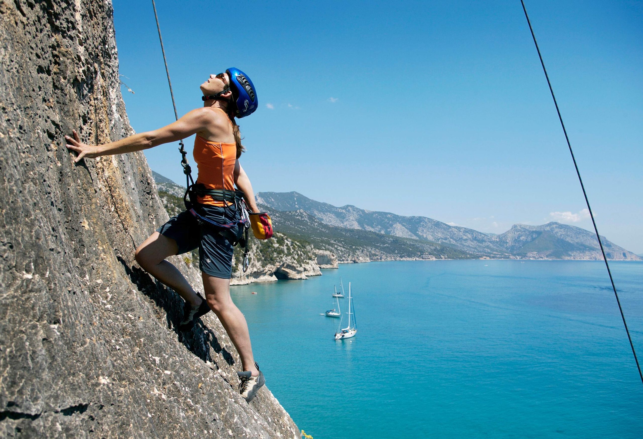 A fearless climber, wearing a helmet and connected to cables, scales a cliff overlooking the awe-inspiring bay below.