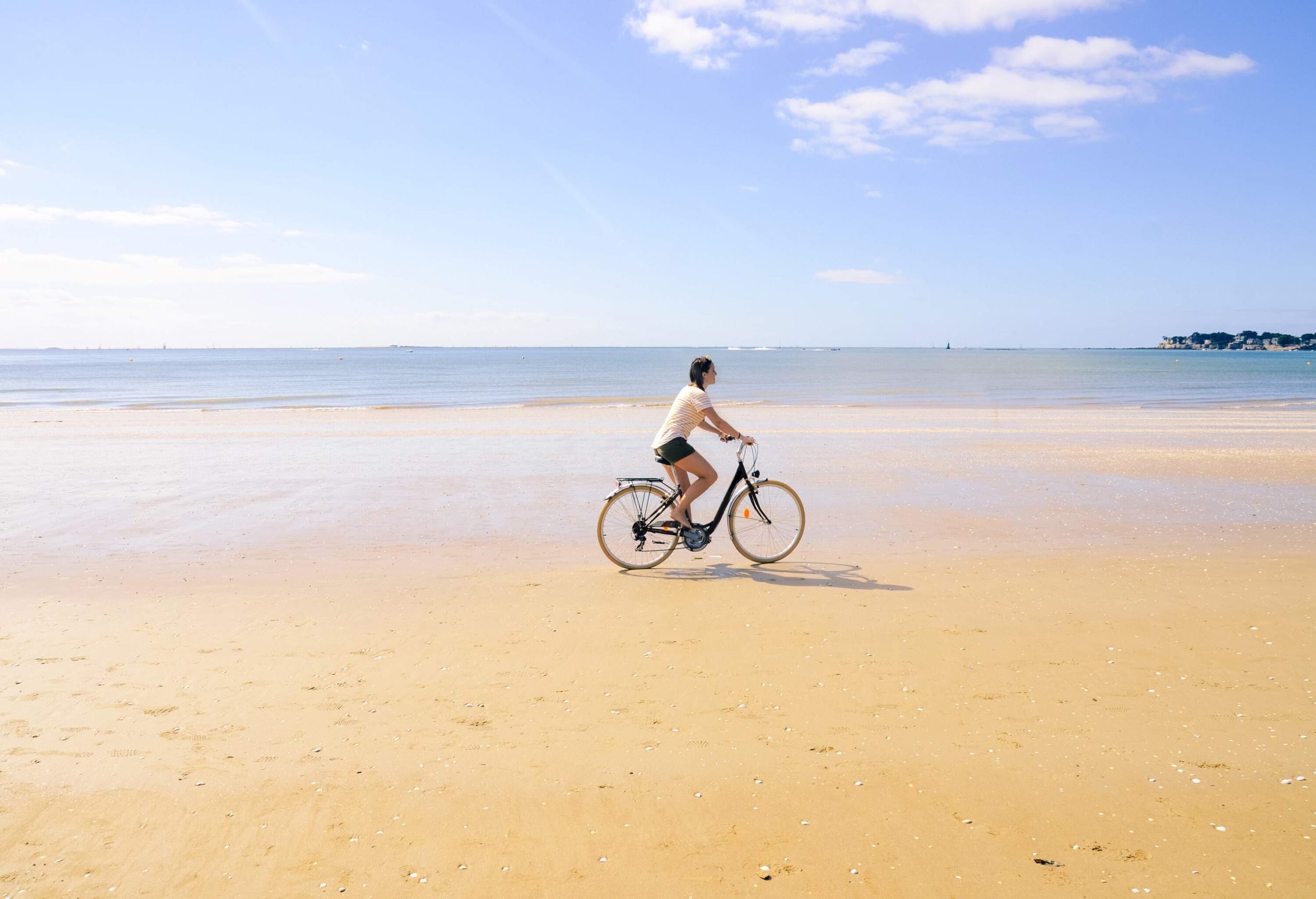 A girl is riding a bike on golden sand with a view of the calm ocean in the background.
