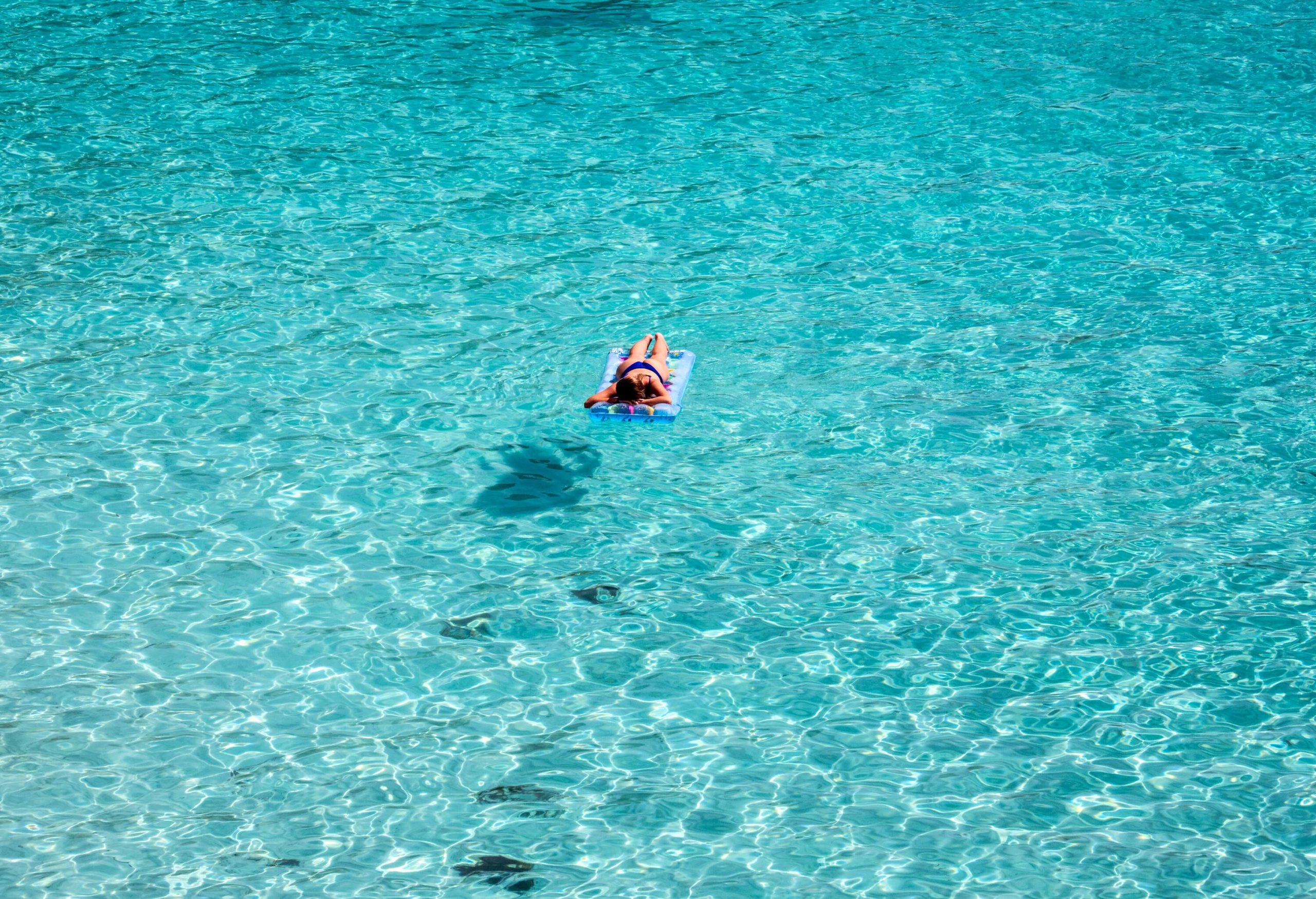 A woman in a blue bikini lying flat on her front facing down in an inflatable floating on a clear turquoise water.