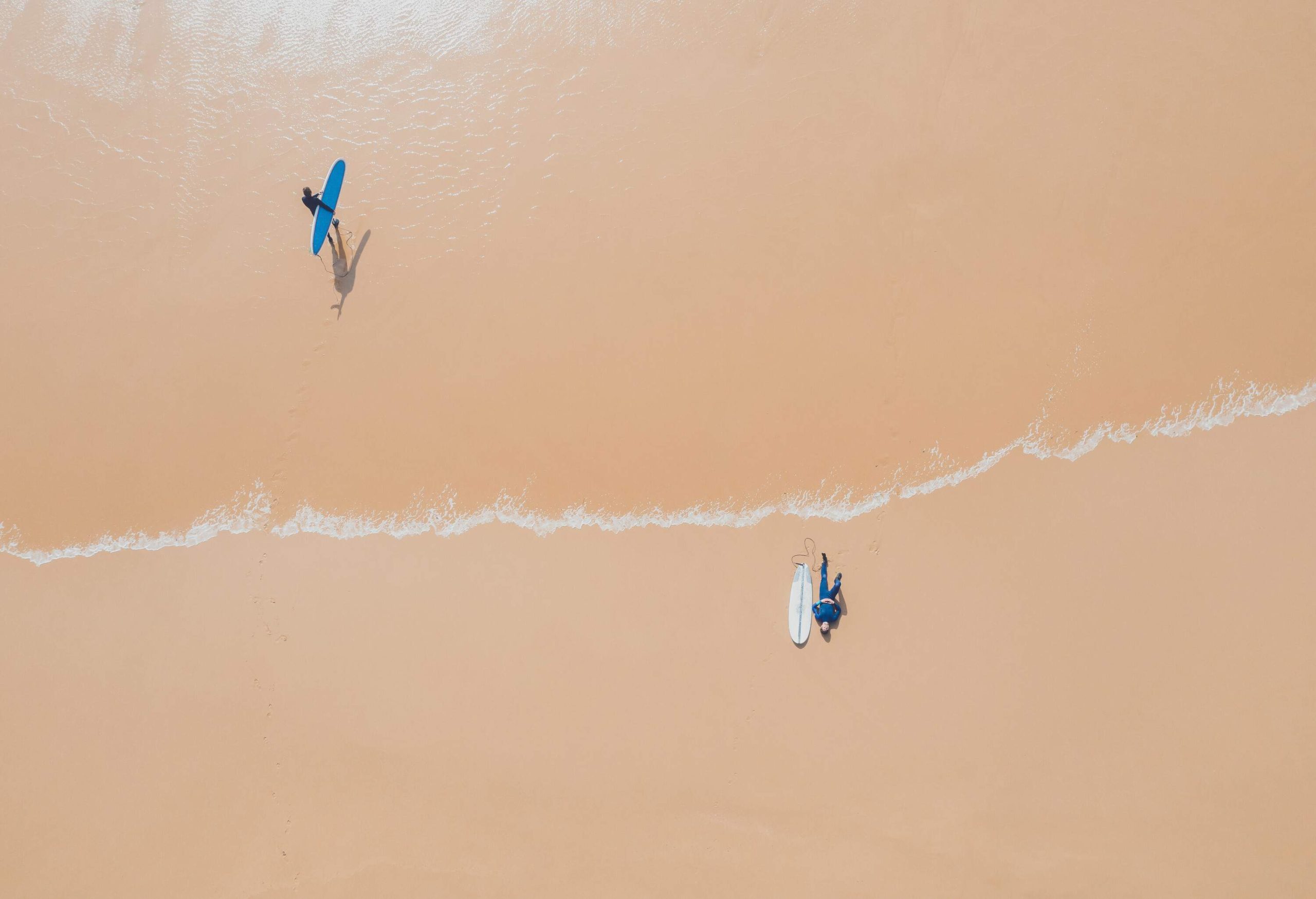 Surfers populate the beach, with one lounging casually beside their surfboard while another strides confidently across the sand, surfboard in tow, ready to conquer the waves.